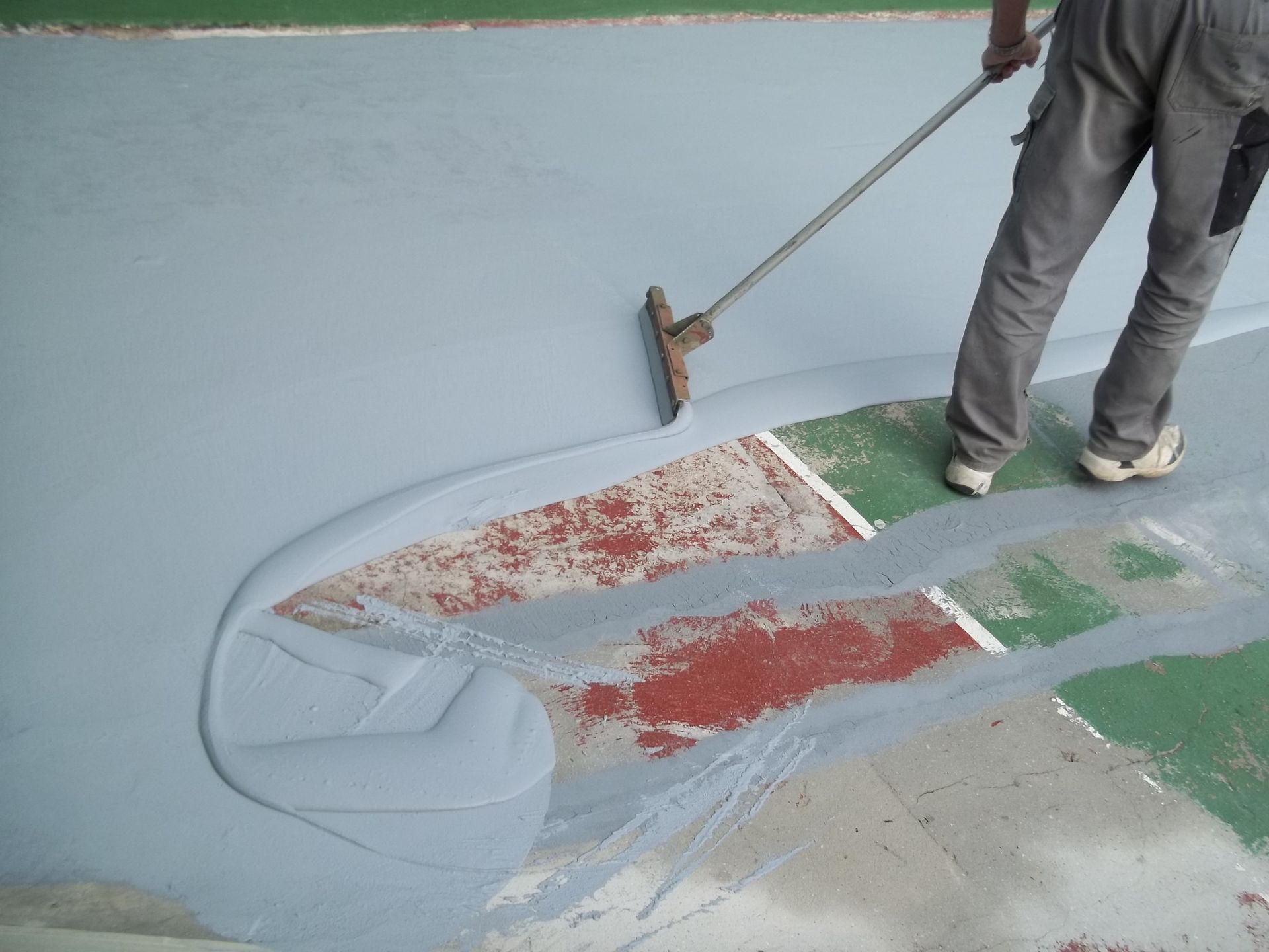 Une personne applique de la peinture grise sur une surface à l'aide d'une raclette. Des zones rouges et vertes sont visibles.