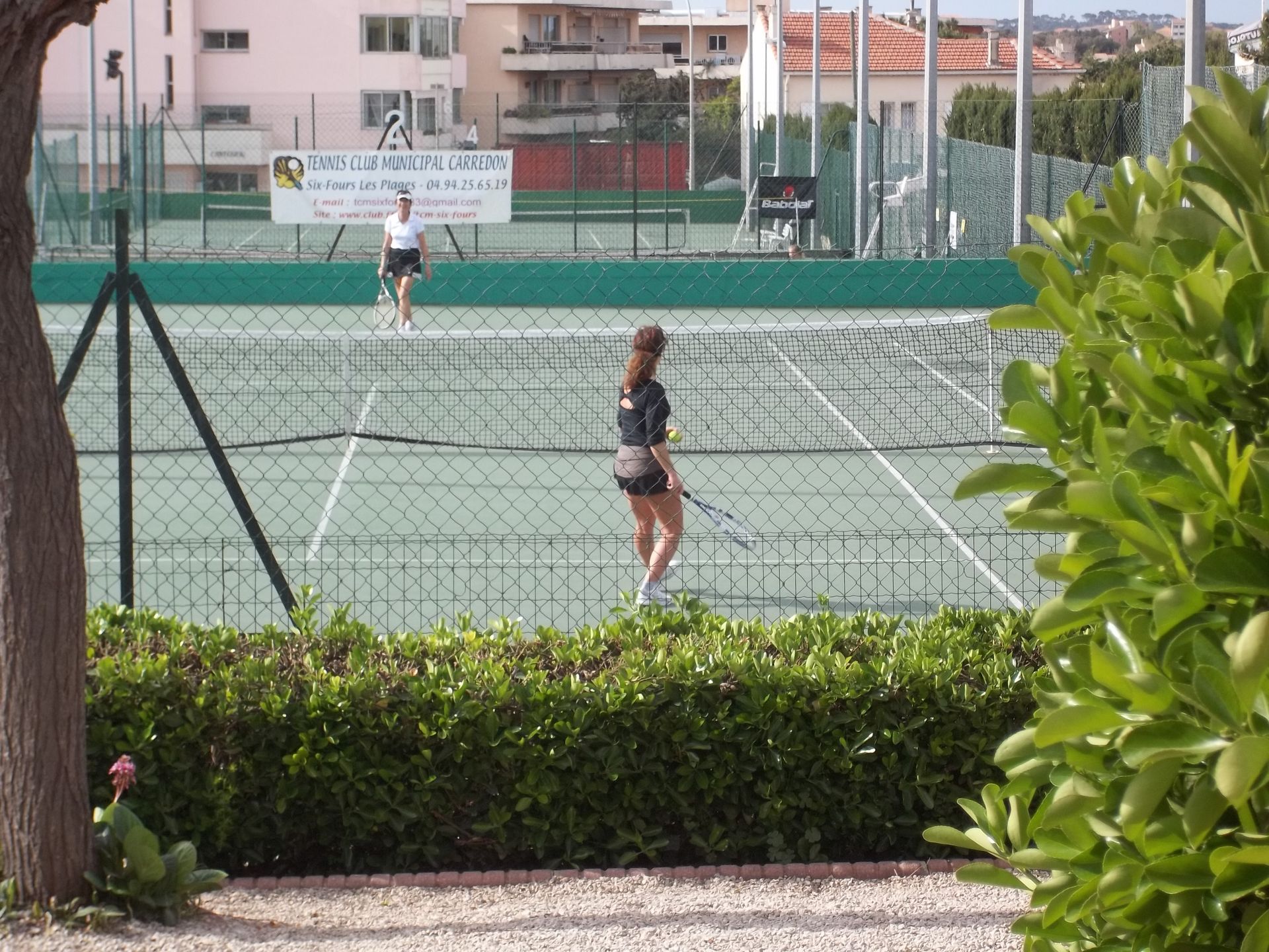 Deux personnes jouent au tennis sur un court vert, une clôture et des bâtiments en arrière-plan.