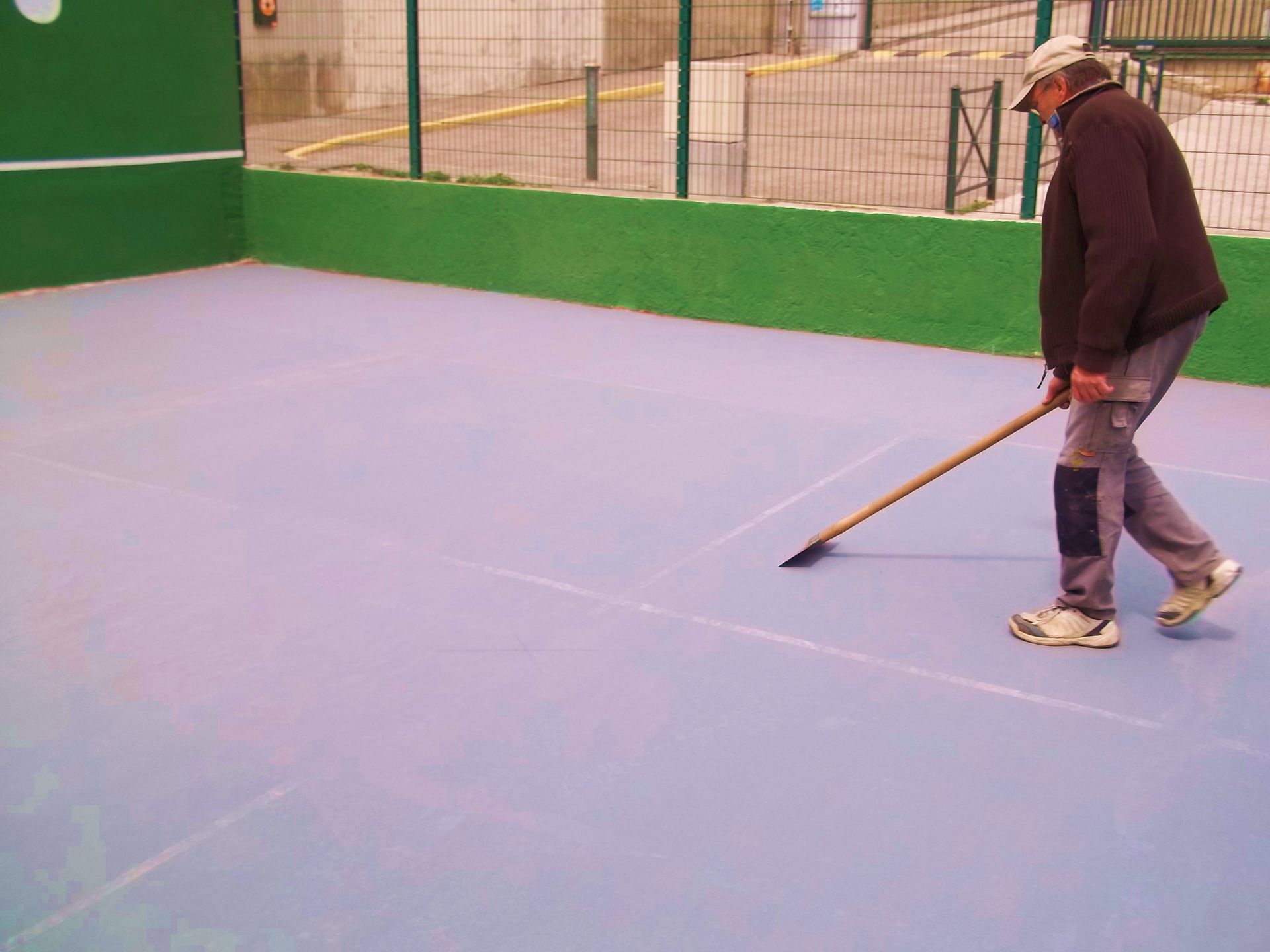 Un homme trace des lignes sur un terrain de sport bleu à l'aide d'un outil à long manche ; un mur vert se trouve en arrière-plan.