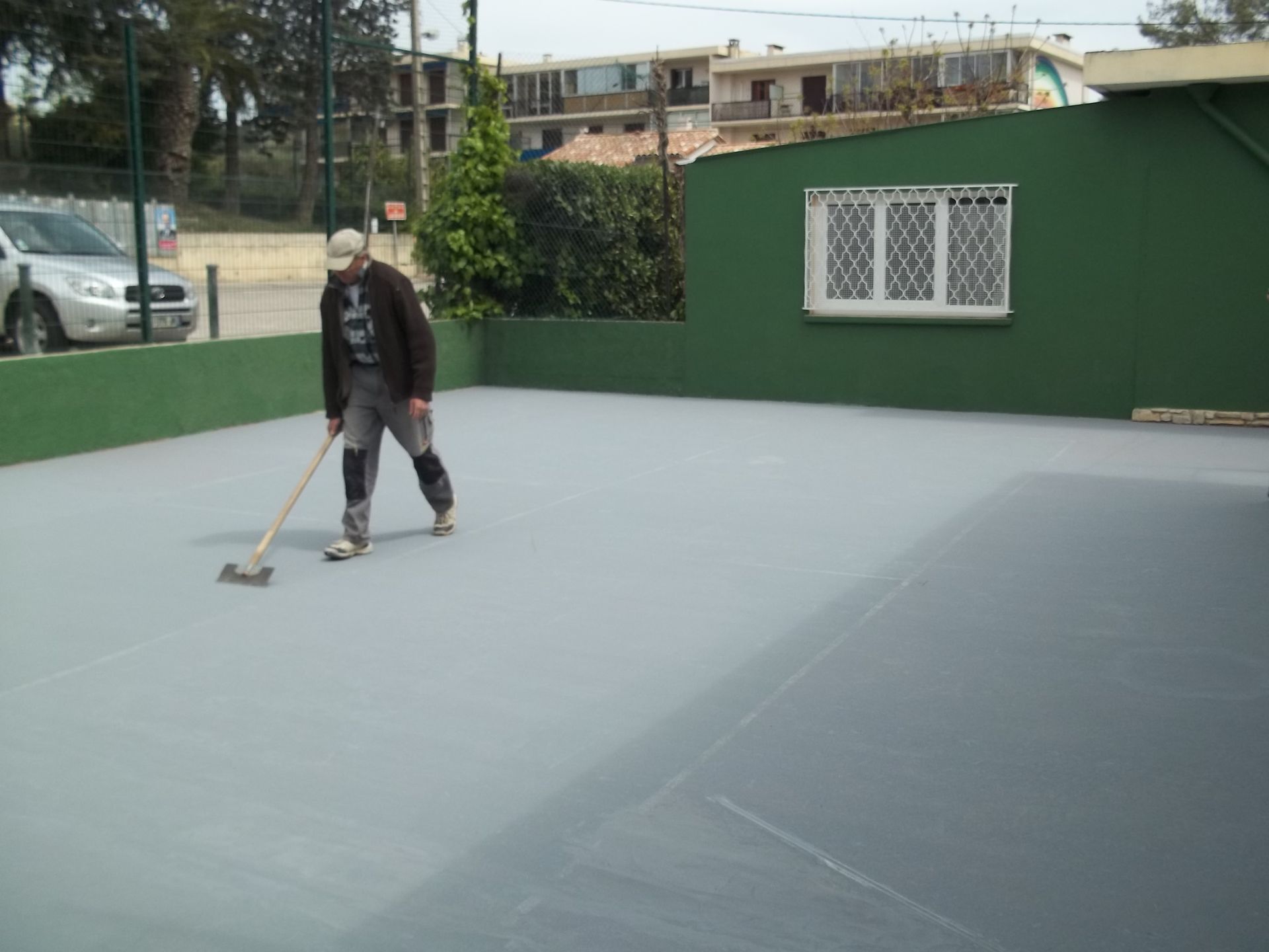 Une personne peint une surface grise à l'extérieur avec un outil à long manche, à côté d'un mur vert, une voiture garée à proximité.