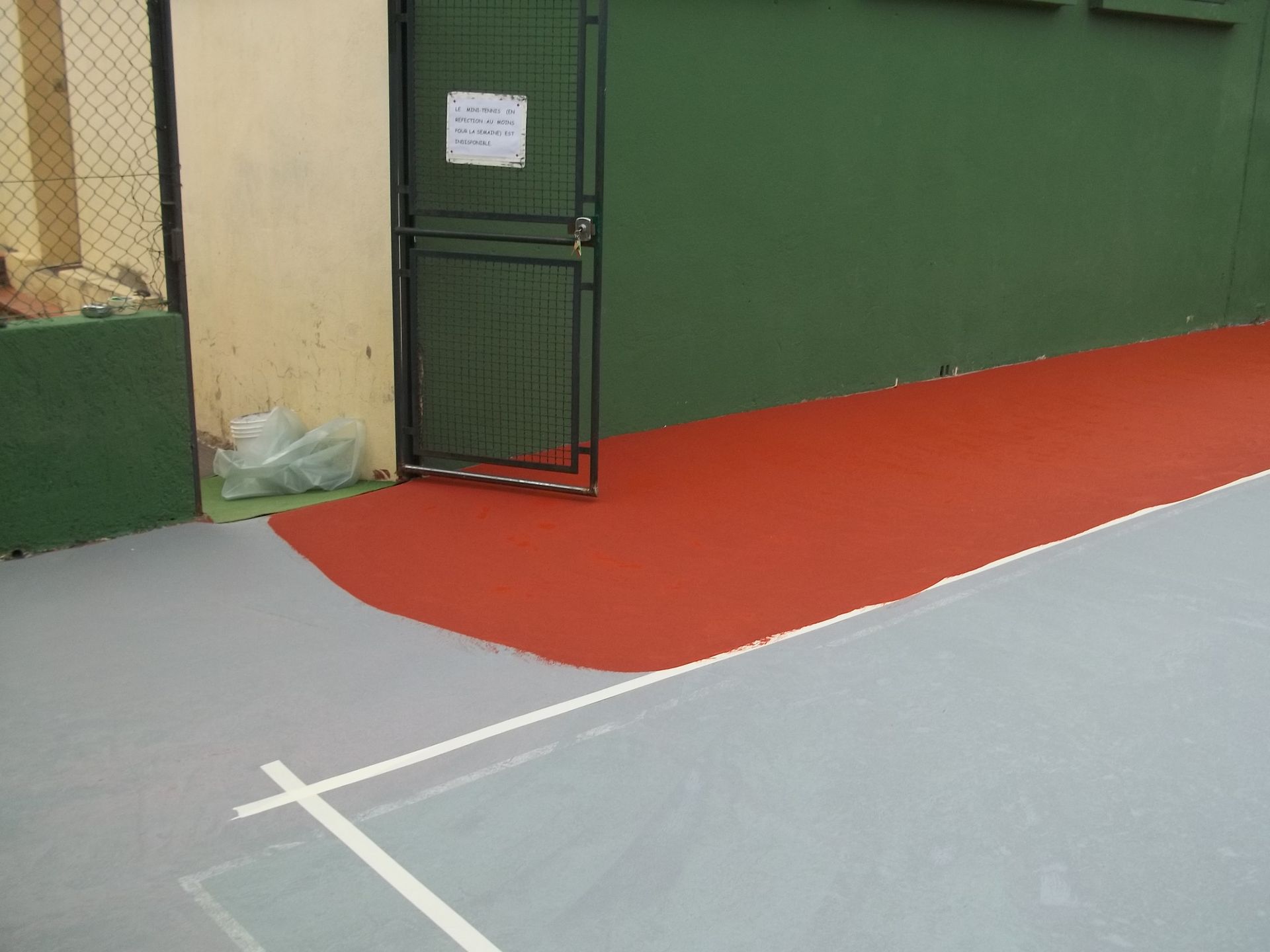 Coin d'un court de tennis avec un sol rouge et gris, un mur vert et un portail noir.
