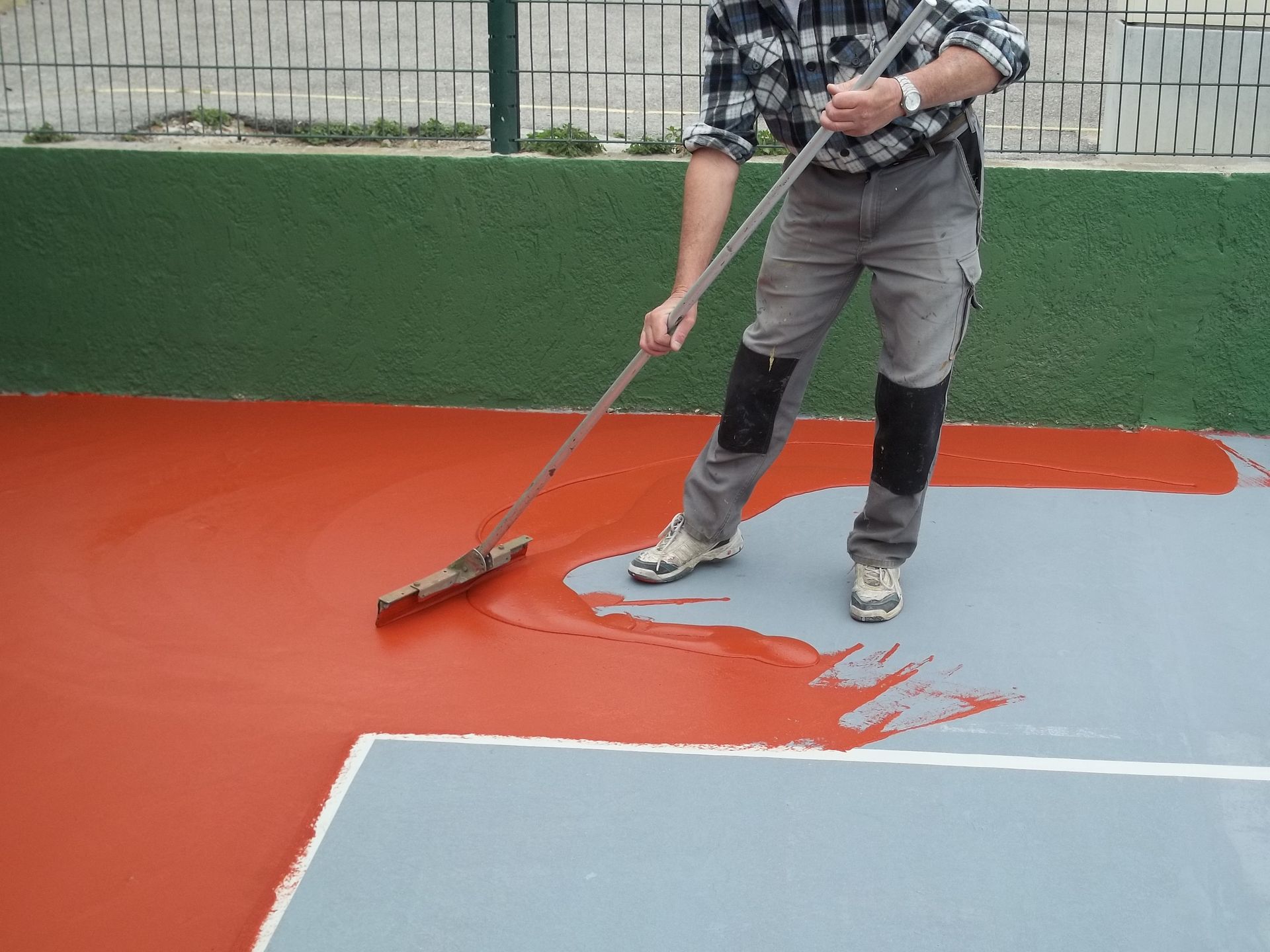 Personne appliquant un enduit rouge sur un court à l'aide d'un outil à long manche.
