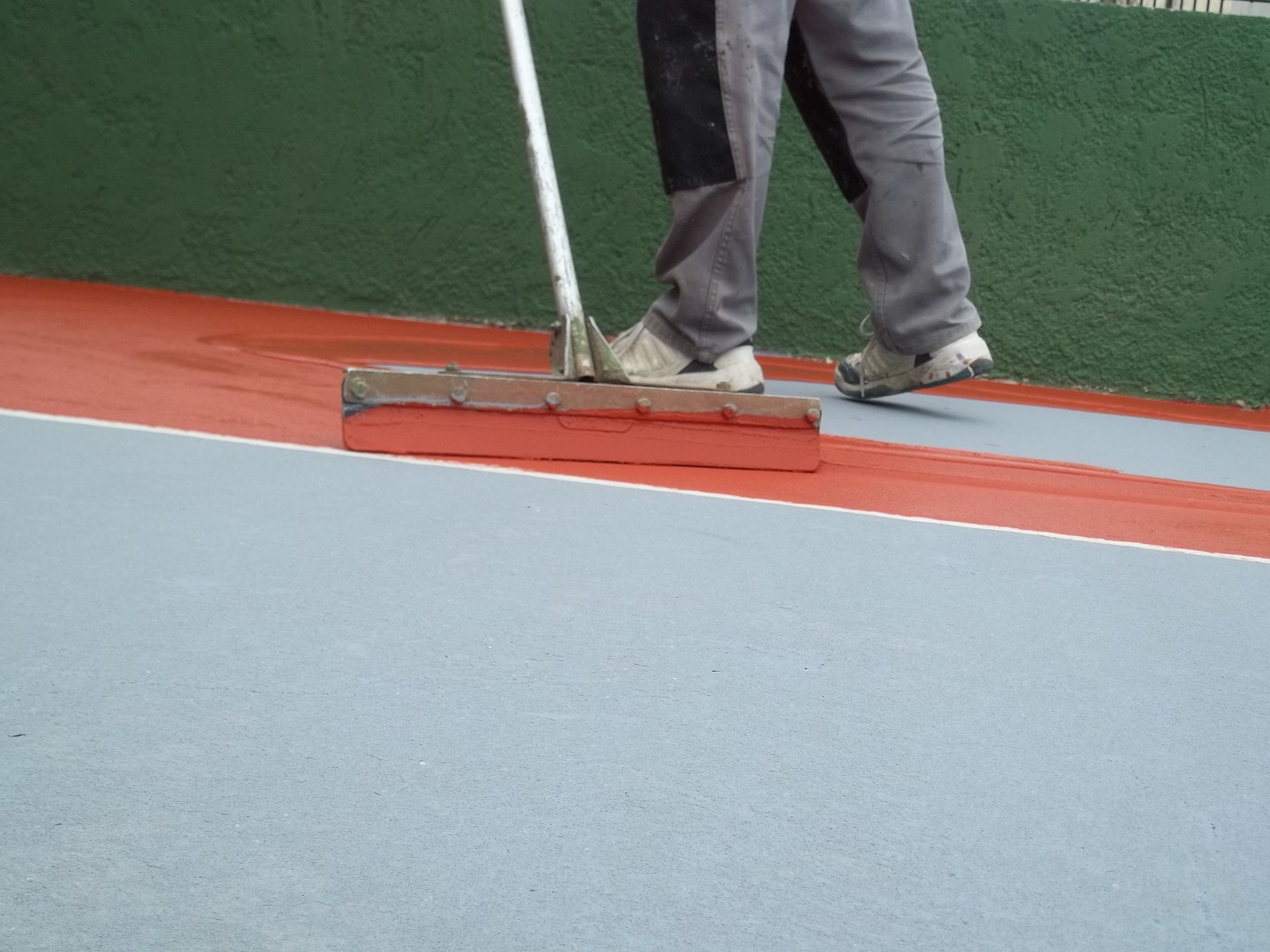 Une personne utilise une raclette pour appliquer un revêtement rouge sur une surface bleue, faisant partie d'un terrain de sport.