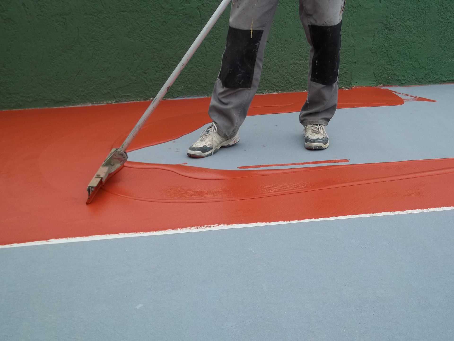 Une personne applique de la peinture rouge sur une surface grise à l'aide d'un outil à long manche ; une ligne blanche sépare les couleurs.