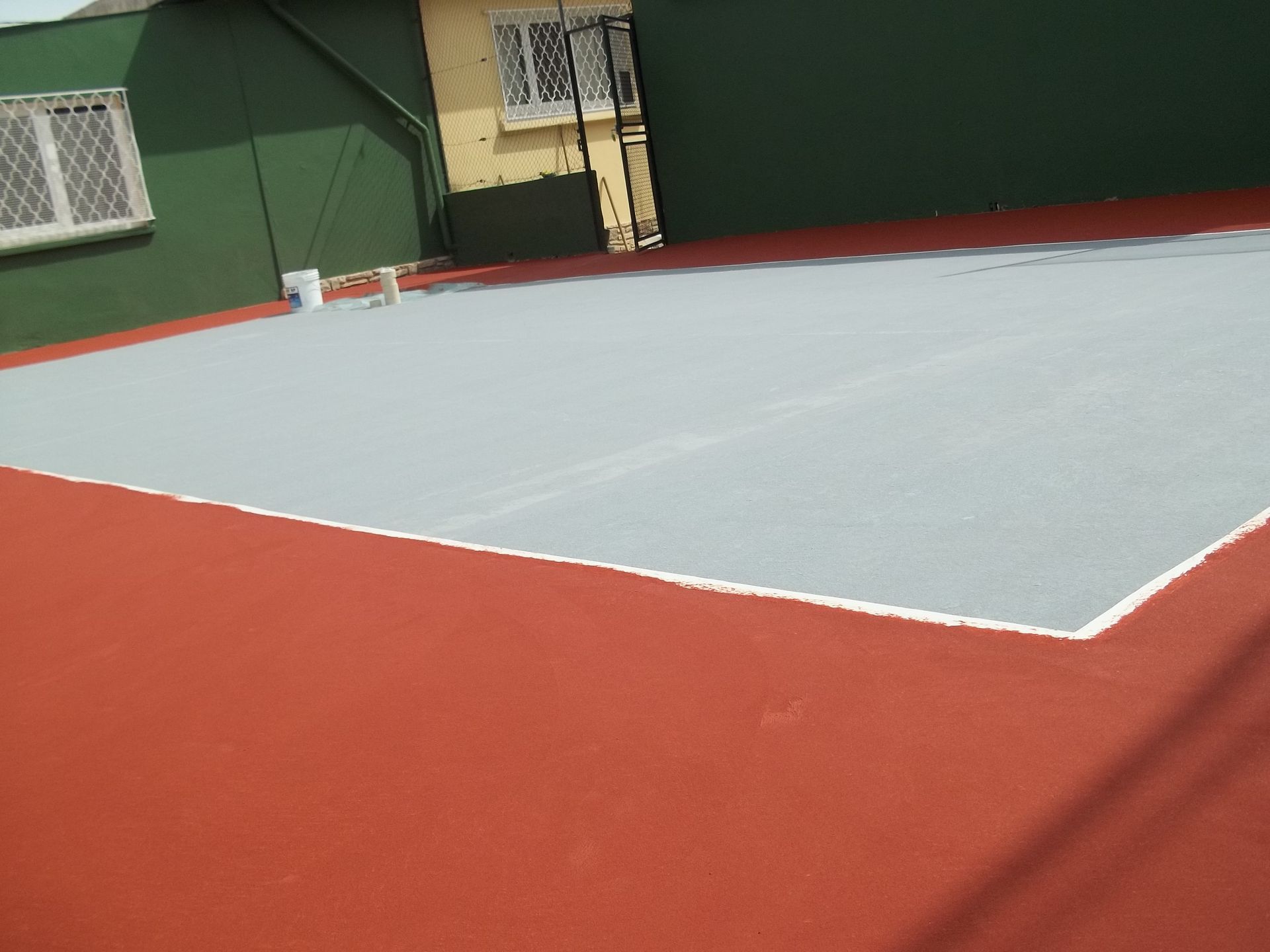 Terrain de basket extérieur rouge et gris avec un bâtiment vert et jaune en arrière-plan.