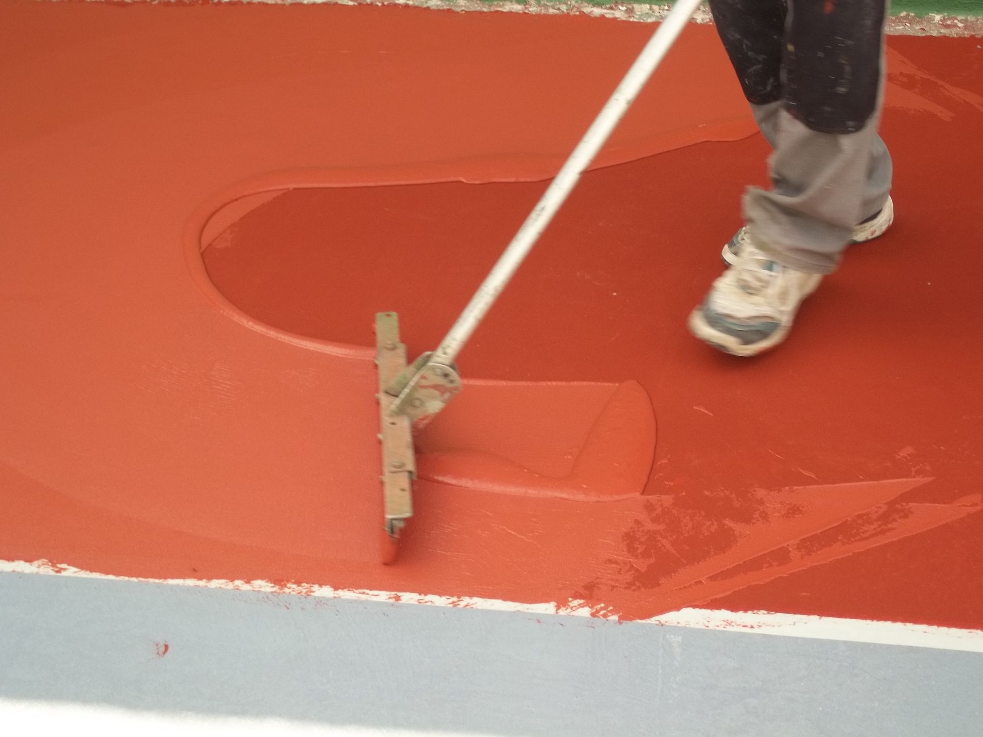 Personne utilisant un grattoir à sol pour appliquer un revêtement rouge sur une surface grise.