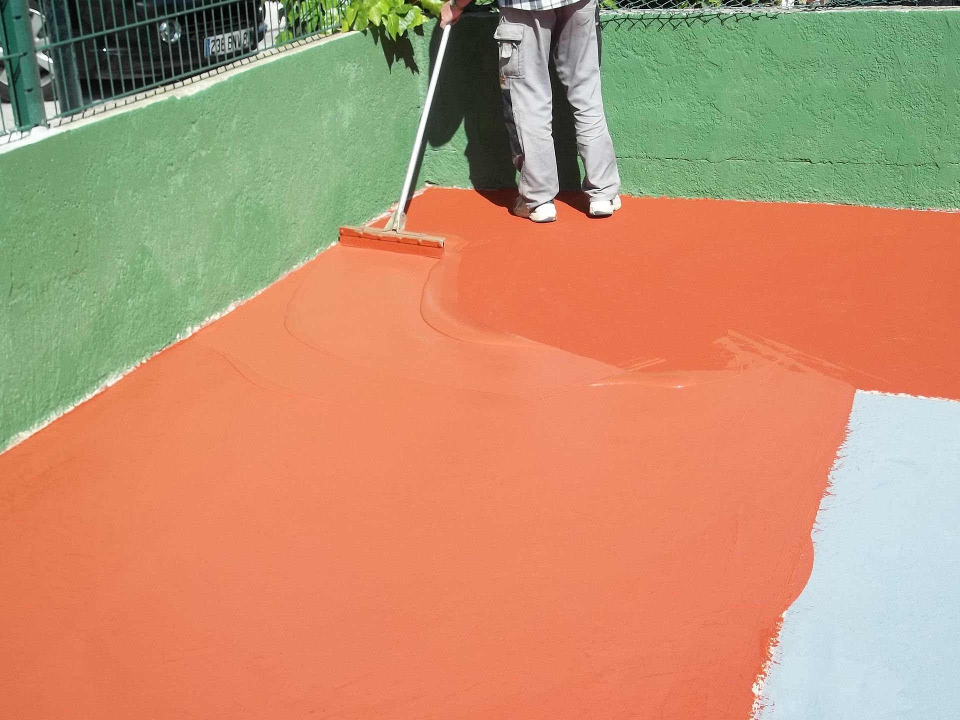 Une personne applique un revêtement rouge sur une surface en béton à l'aide d'une raclette à long manche ; des surfaces vertes et bleues sont également visibles.