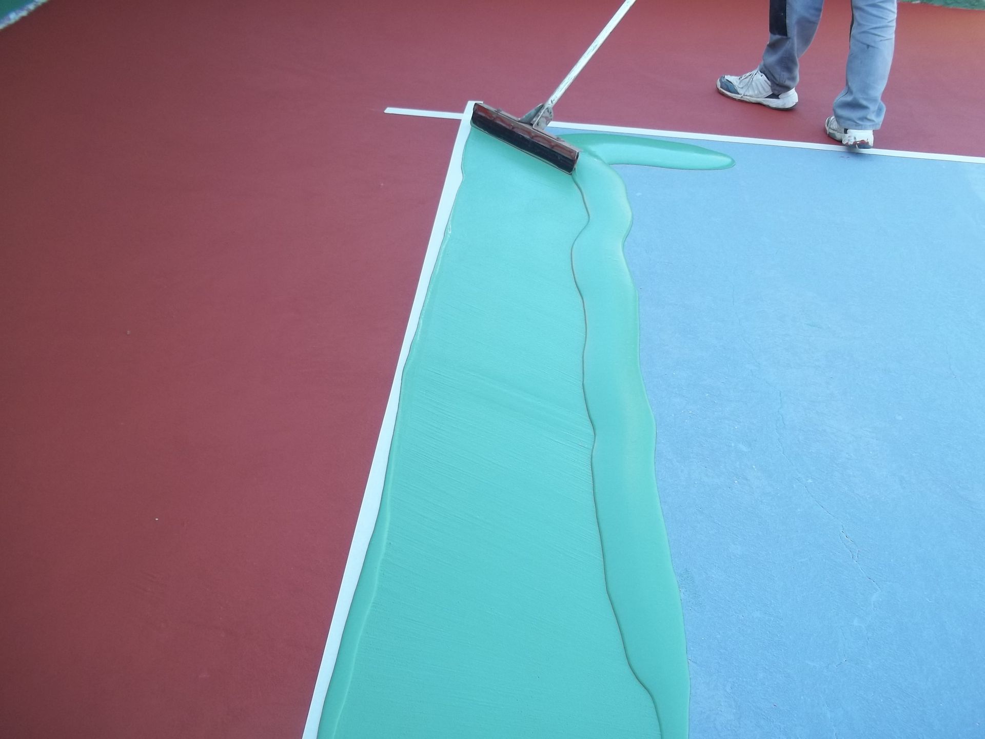 Une personne utilise un rouleau pour appliquer un revêtement vert sur un terrain bleu adjacent à une zone rouge, créant ainsi une bordure.