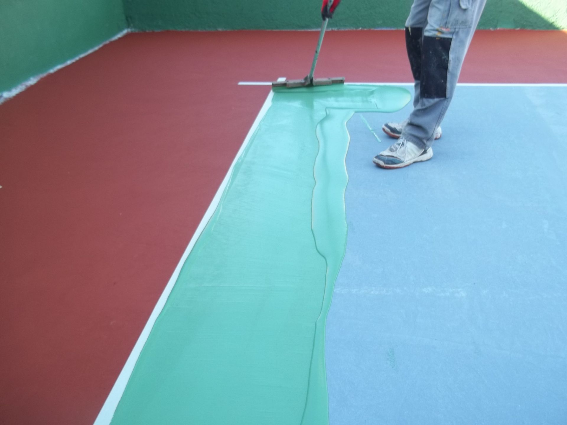 Une personne applique de la peinture verte sur un court de tennis bleu, près d'une zone rouge.