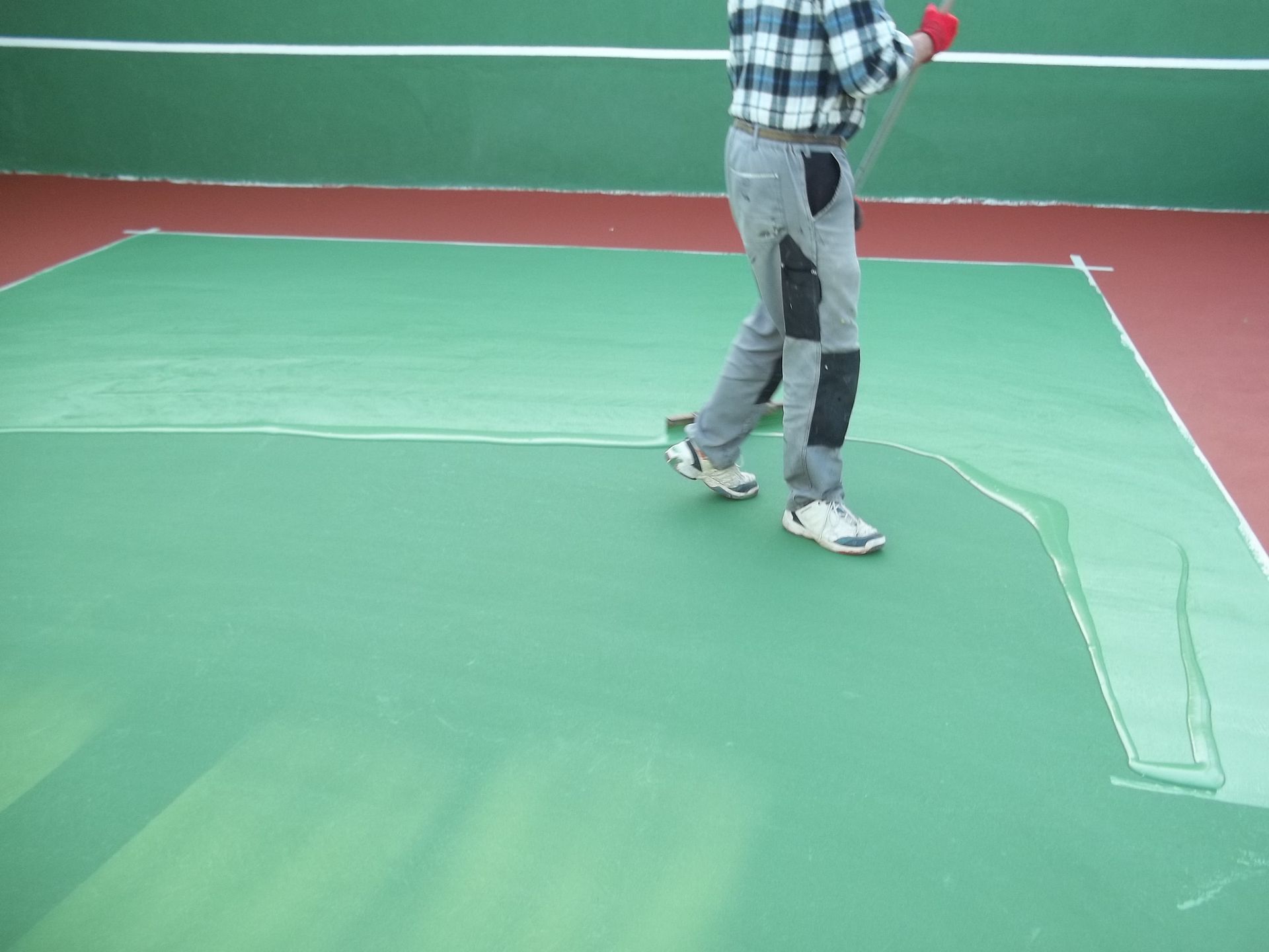 Une personne peint un court de tennis vert avec un rouleau à long manche, court rouge/vert avec des lignes blanches.