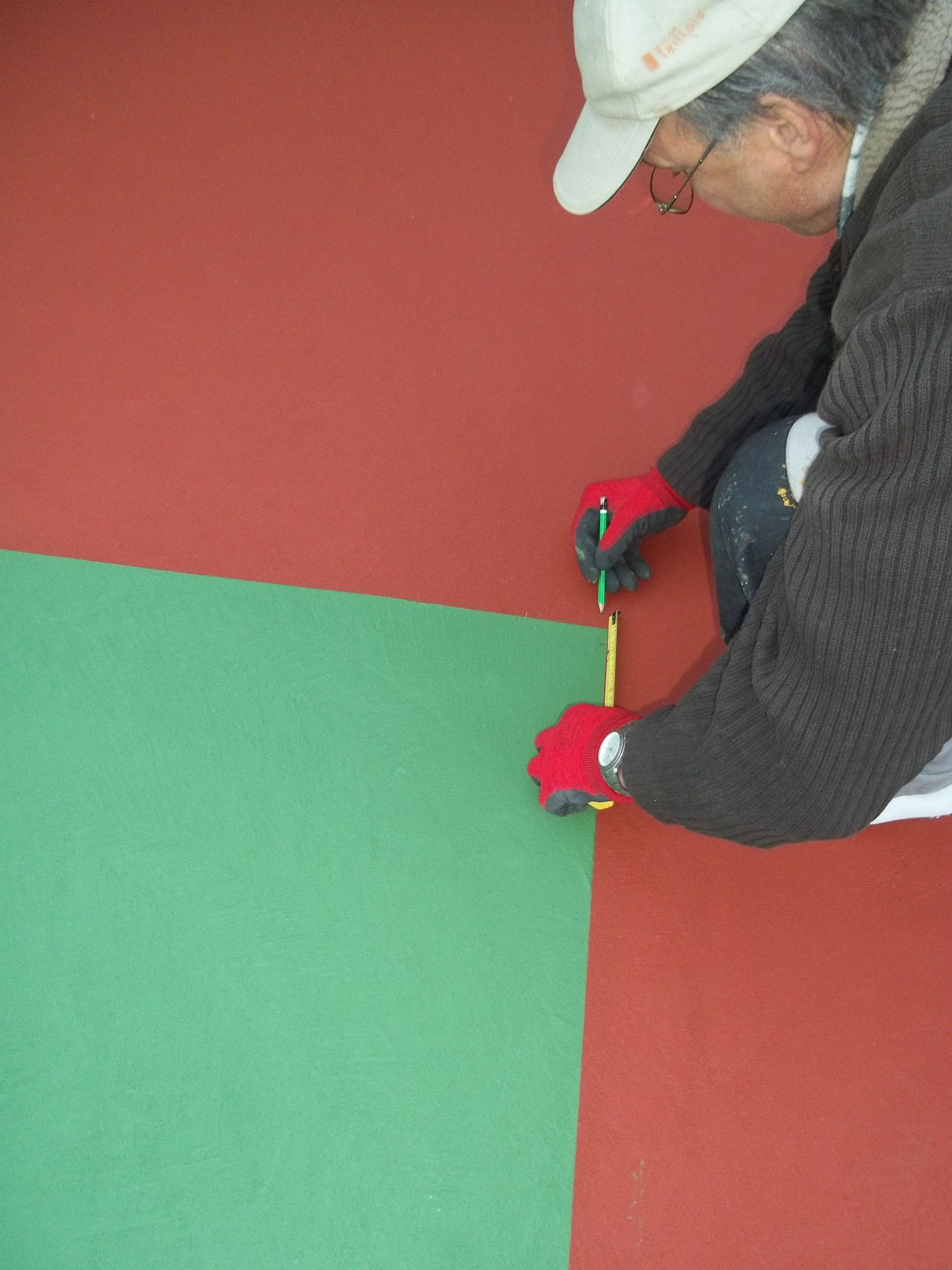 Une personne portant des gants et un chapeau mesure un carré vert contre un mur rouge, à l'aide d'un outil de mesure jaune.