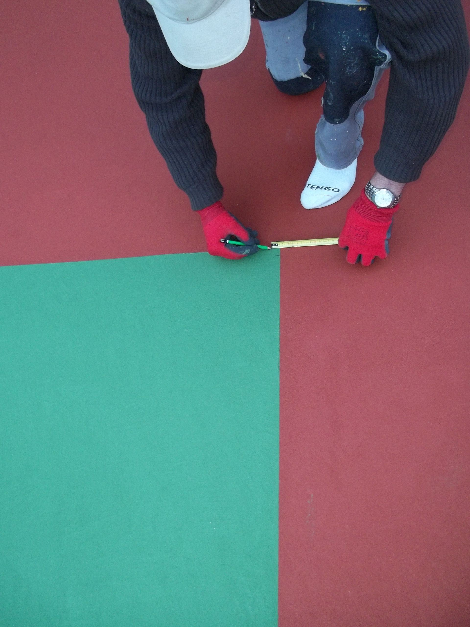 Une personne coupant un carreau de sol vert à côté d'un carreau rouge, portant des gants rouges, une casquette et tenant un mètre ruban.