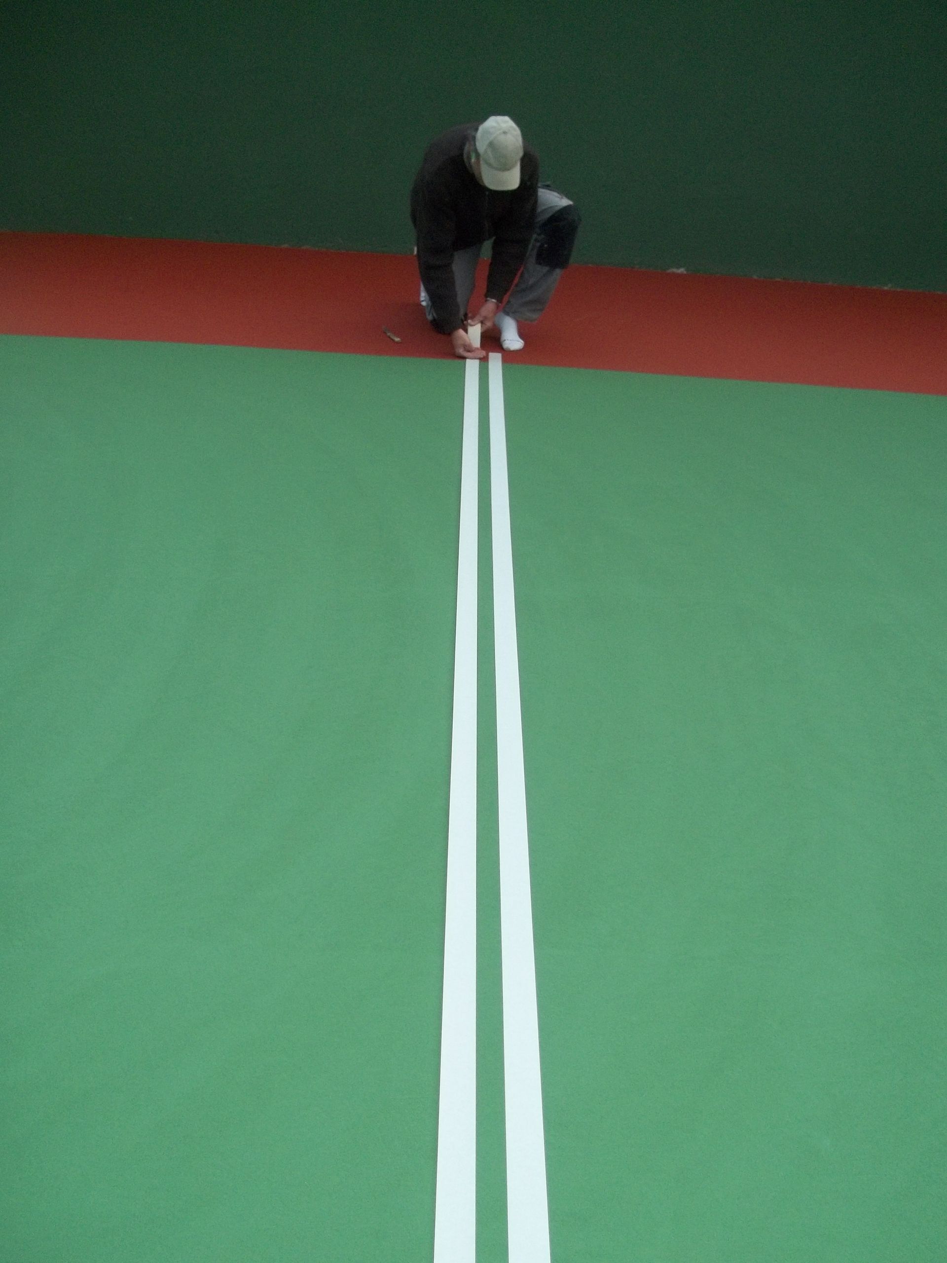 Personne agenouillée, peignant des lignes blanches sur une surface verte. Arrière-plan rouge et vert foncé.