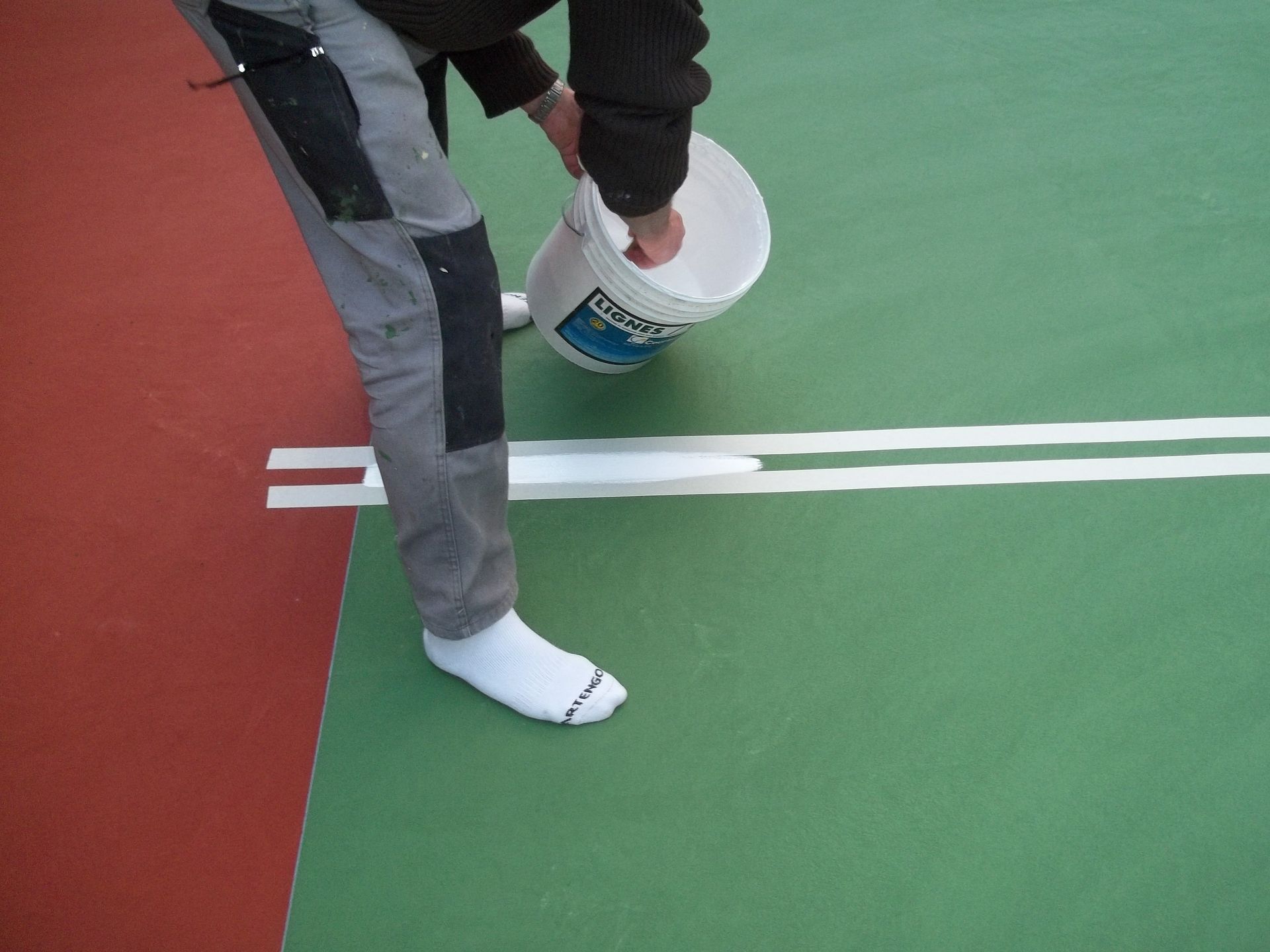Une personne verse de la peinture blanche d'un seau sur un terrain de sport vert, en suivant les lignes tracées au ruban adhésif de masquage.