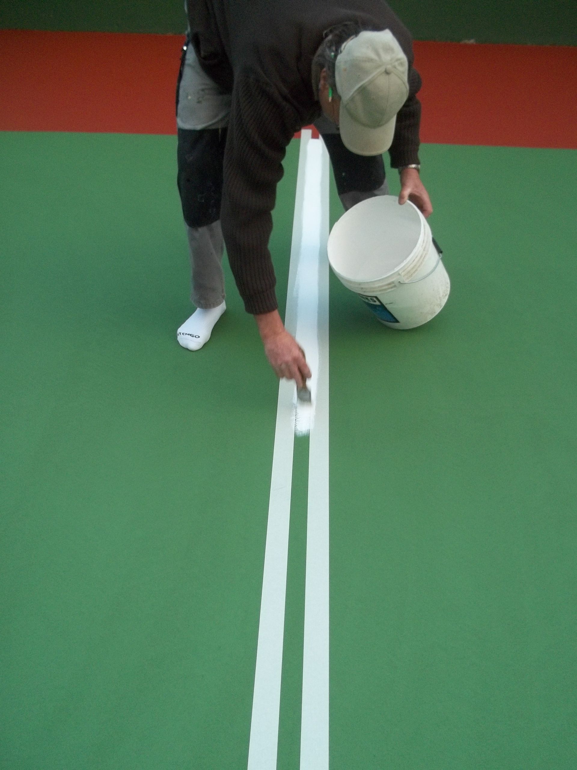 Une personne peignant une ligne blanche sur un court de tennis vert, tenant un seau.
