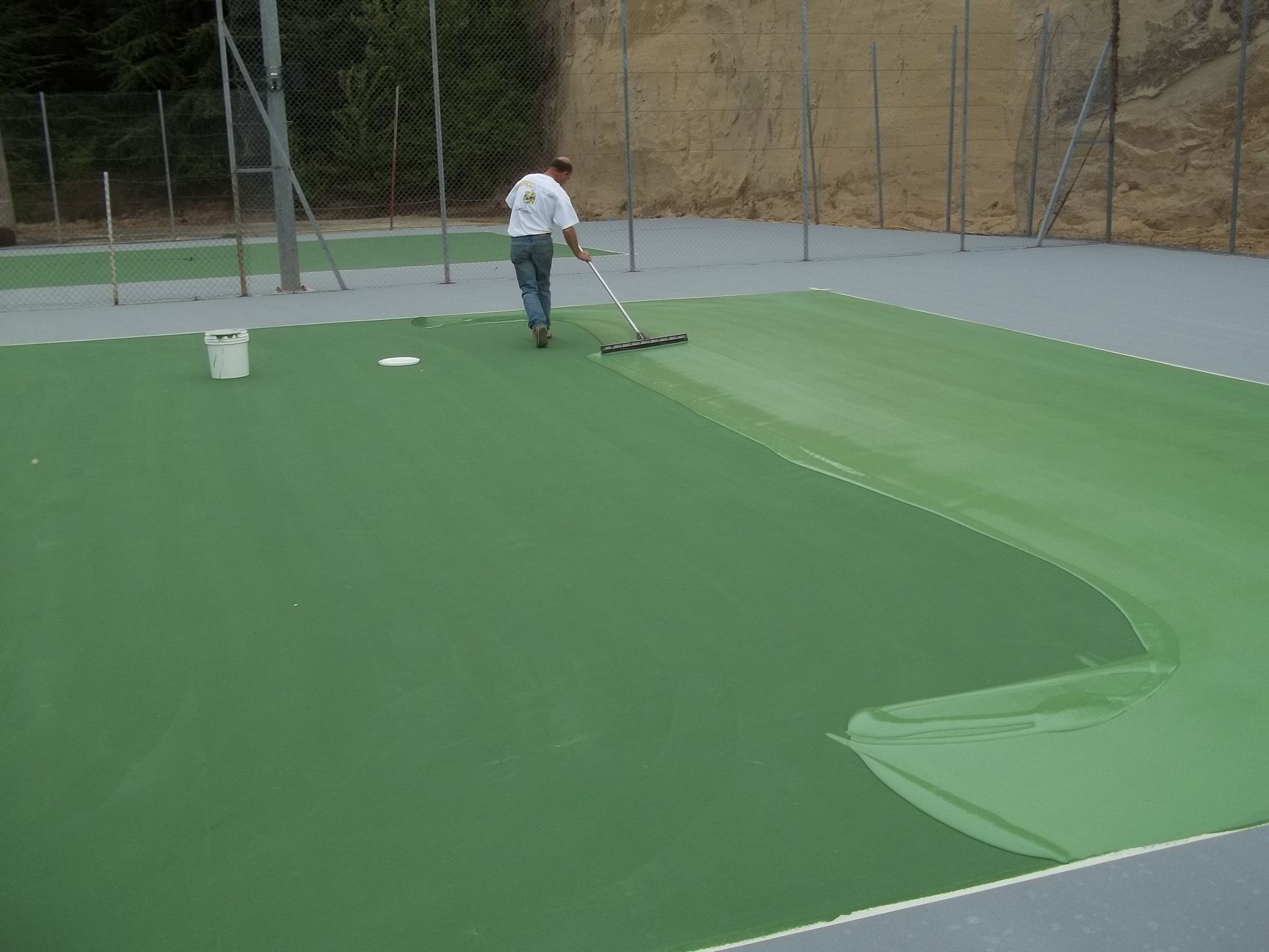 Un homme peint un court de tennis en vert avec un rouleau, à l'extérieur.