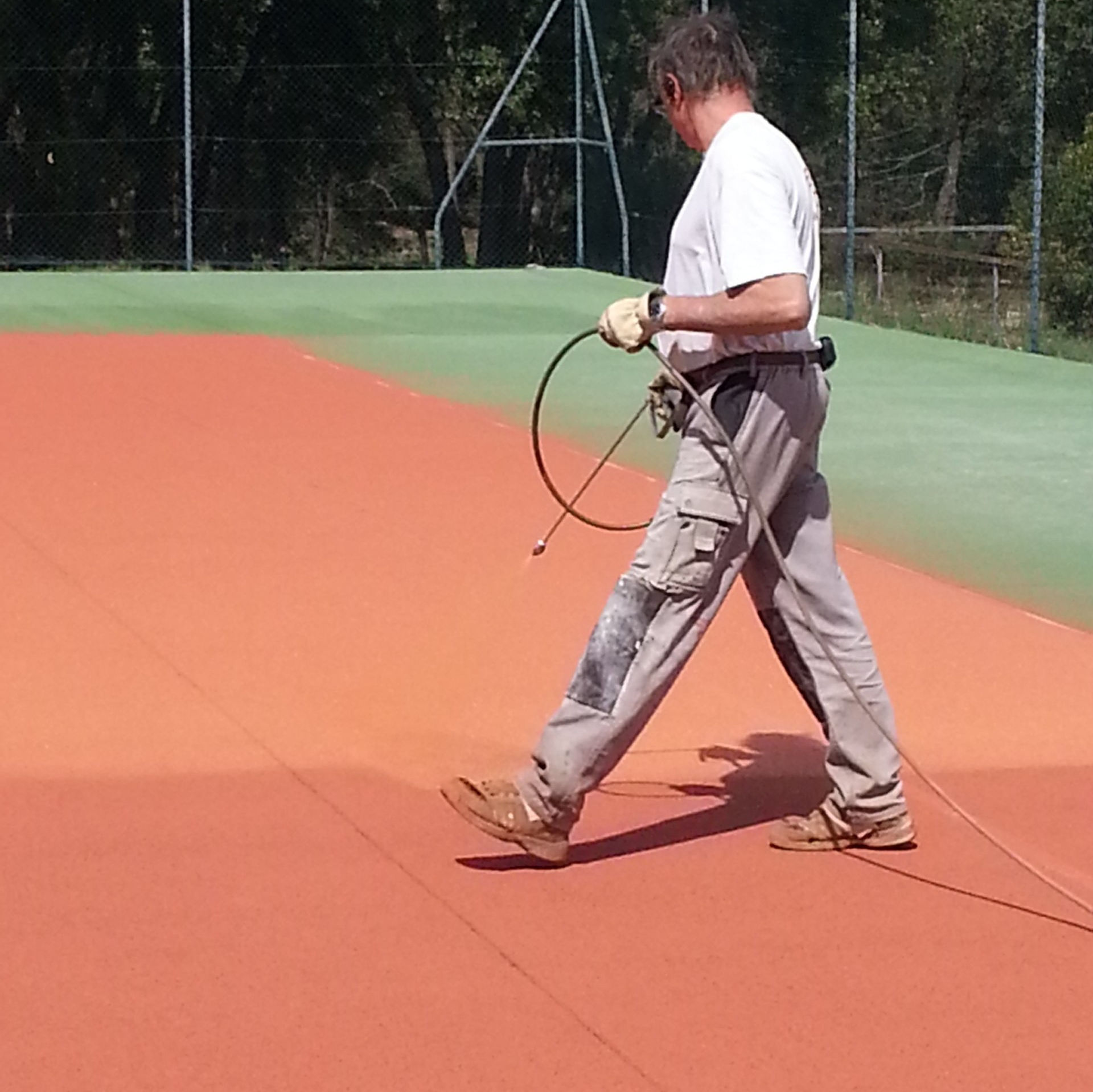 Un homme pulvérise un produit sur un court de tennis avec un revêtement rouge, vêtu d'une chemise blanche, d'un pantalon beige et de gants de travail.