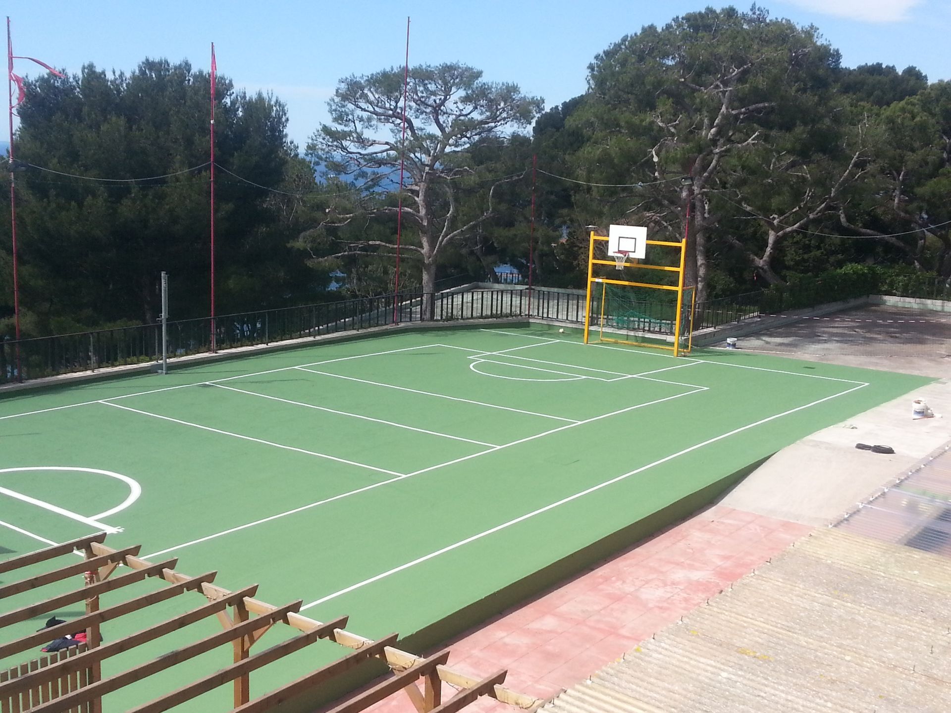 Terrain de basket-ball, surface verte avec lignes blanches, panier jaune, arbres et ciel bleu.