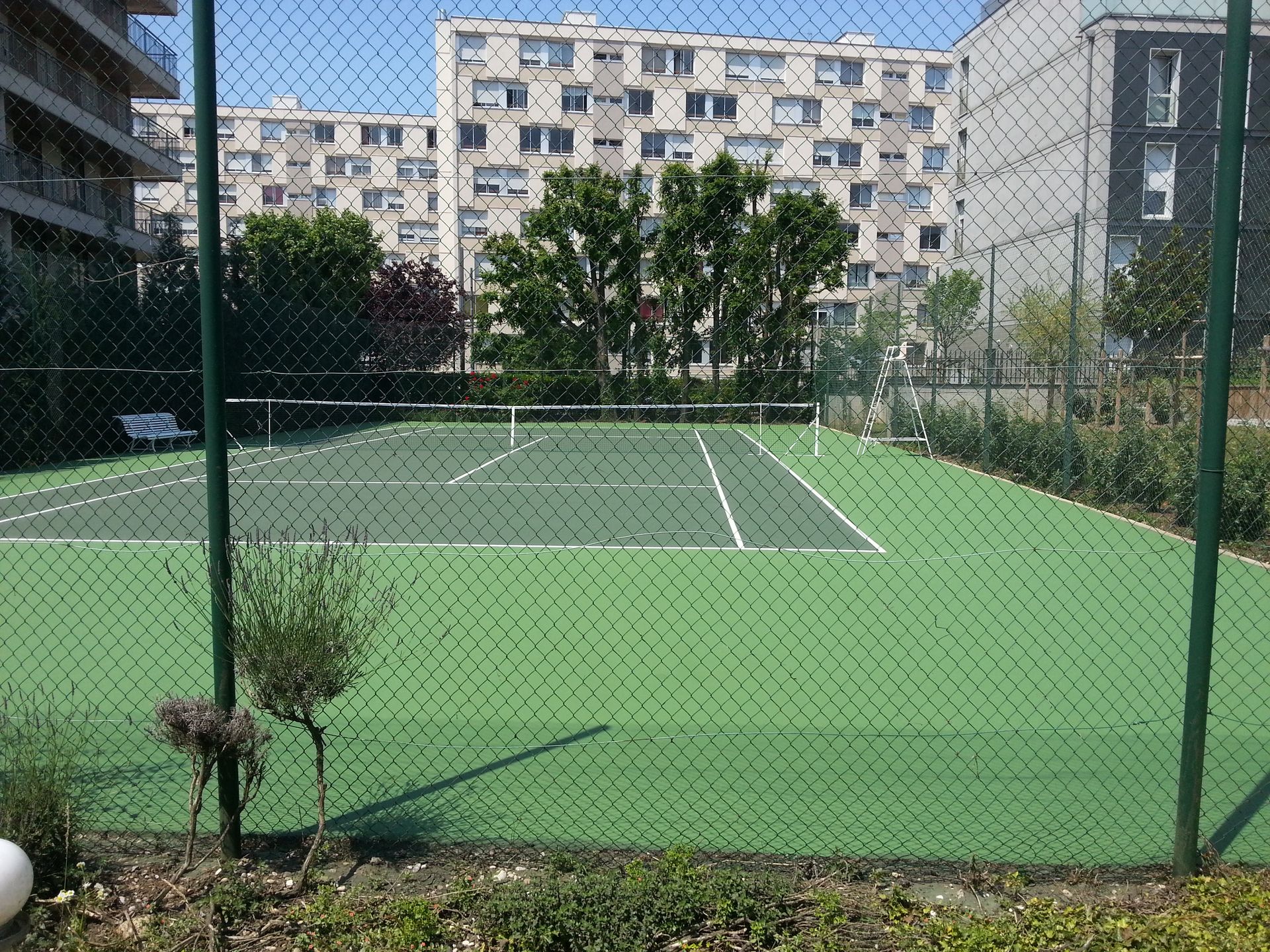 Court de tennis verdoyant derrière une clôture en grillage, avec des immeubles d'appartements en arrière-plan par une journée ensoleillée.