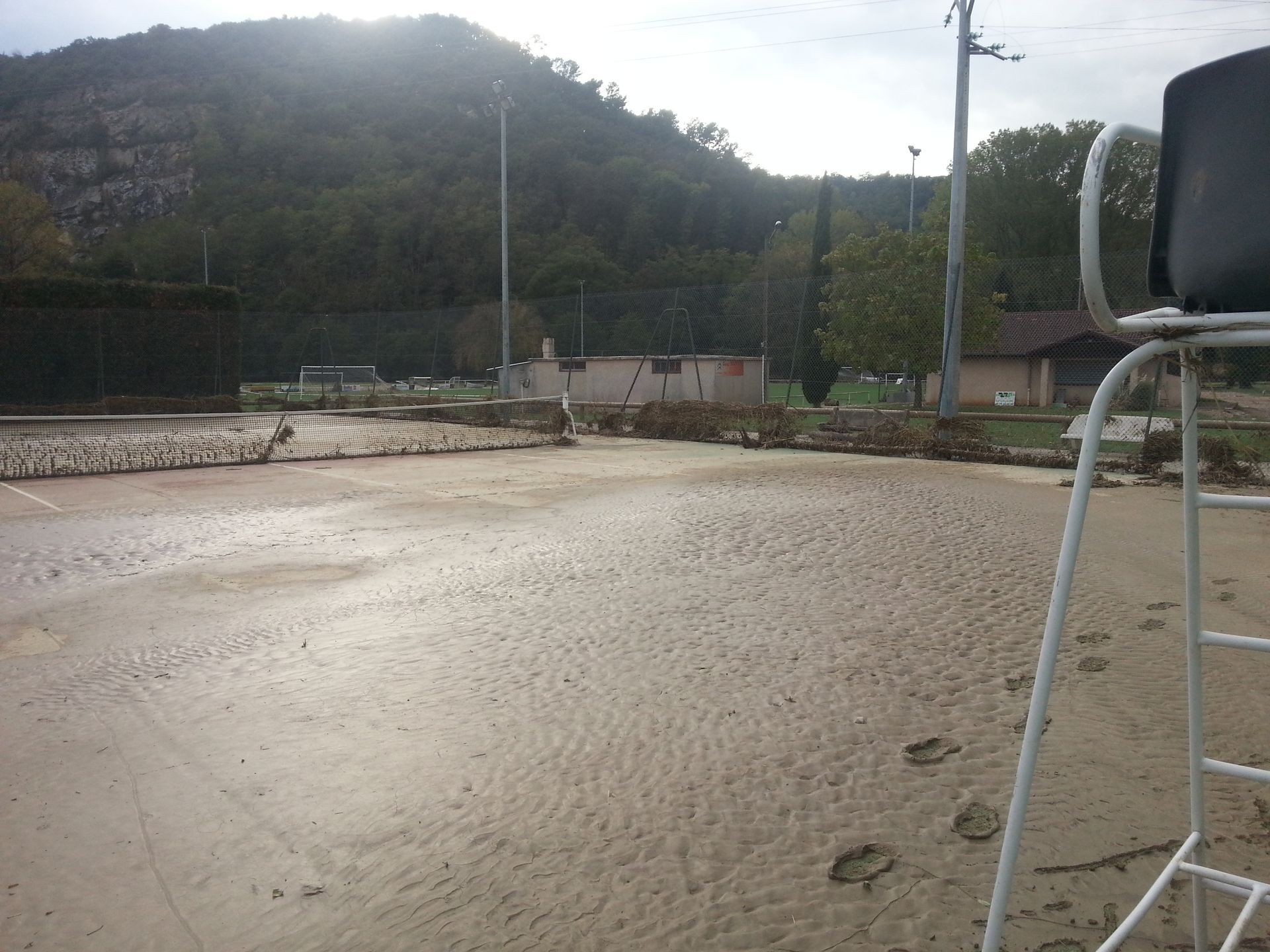 Court de tennis recouvert de boue après une inondation, avec des débris, les bâtiments du court et des arbres en arrière-plan.