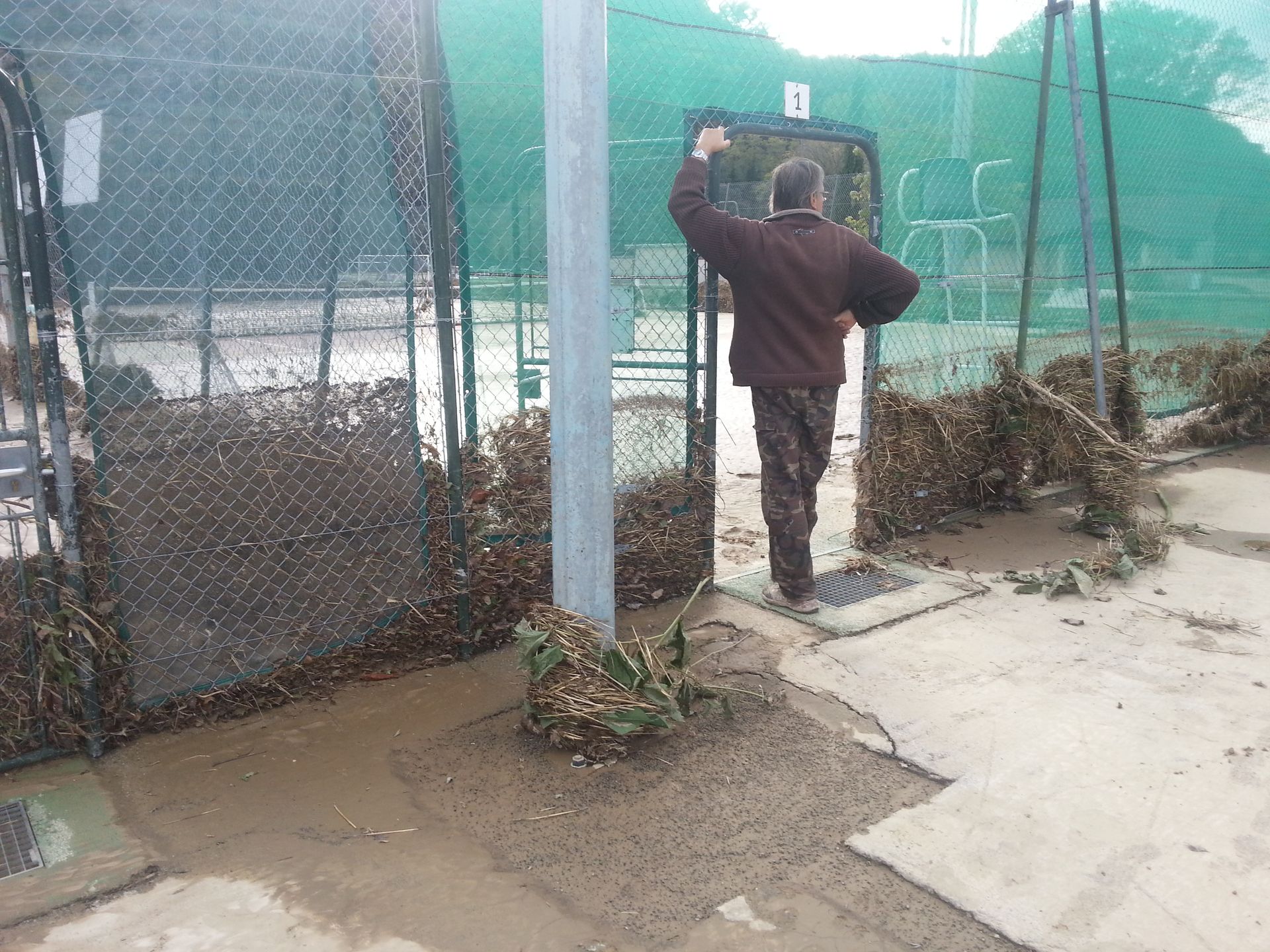 Un homme ouvre un portail. Terrain boueux, clôture en grillage.