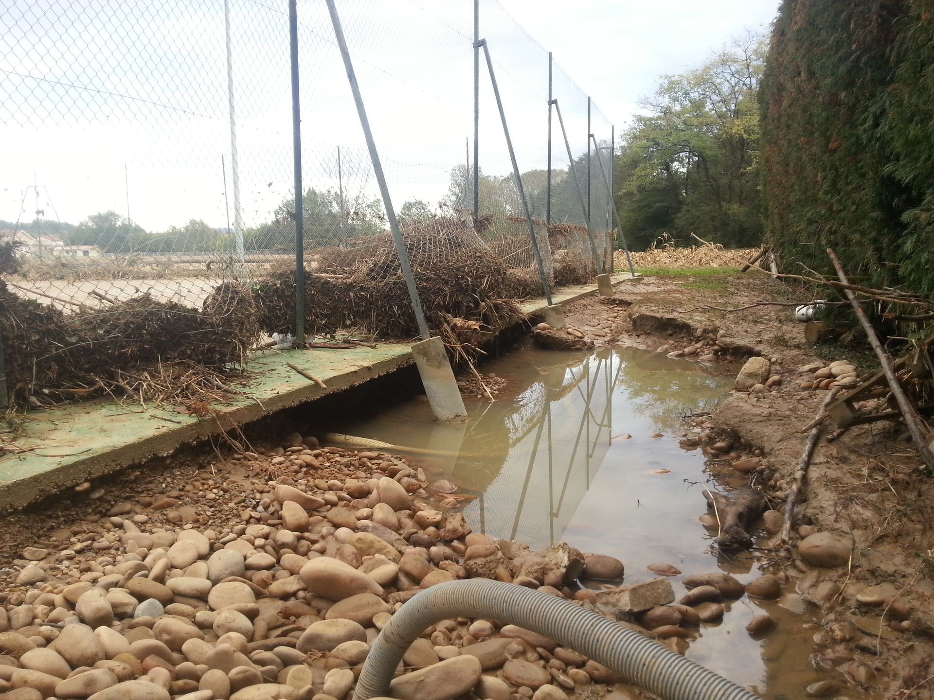 Zone inondée avec des débris, une clôture et de l'eau brunâtre ; ciel couvert en arrière-plan.