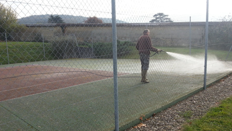 Une personne arrose un court de tennis. Court vert, clôture en grillage, pelouse et colline en arrière-plan.