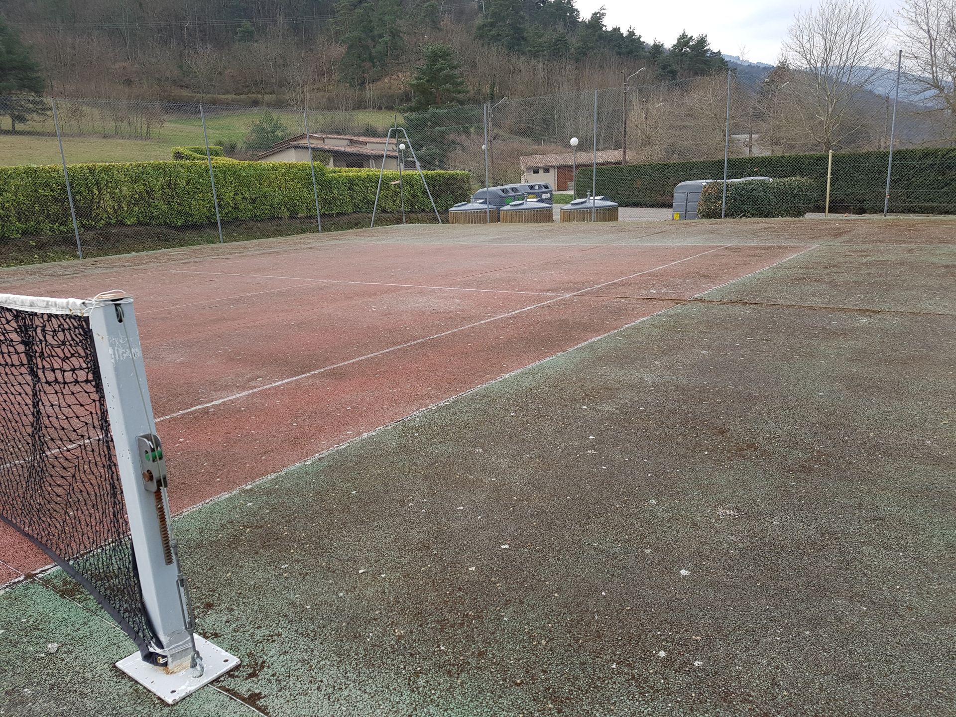 Court de tennis, filet, surface en terre battue rouge, pelouse, arrière-plan d'arbres et de bâtiments bas.