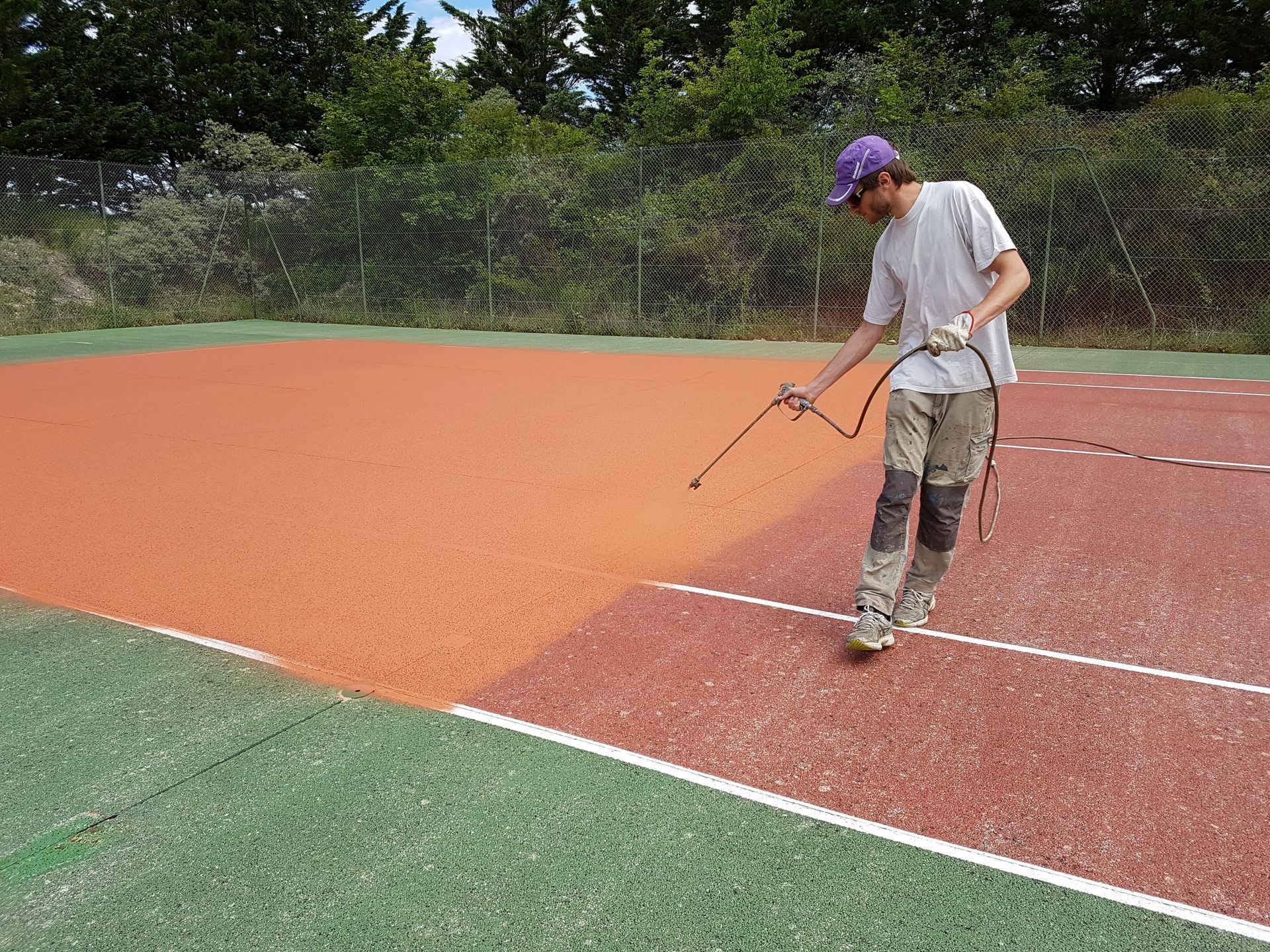 Une personne pulvérise un enduit protecteur orange sur un terrain multisport. Le gazon synthétique est vert au premier plan, avec des lignes blanches et un filet noir.