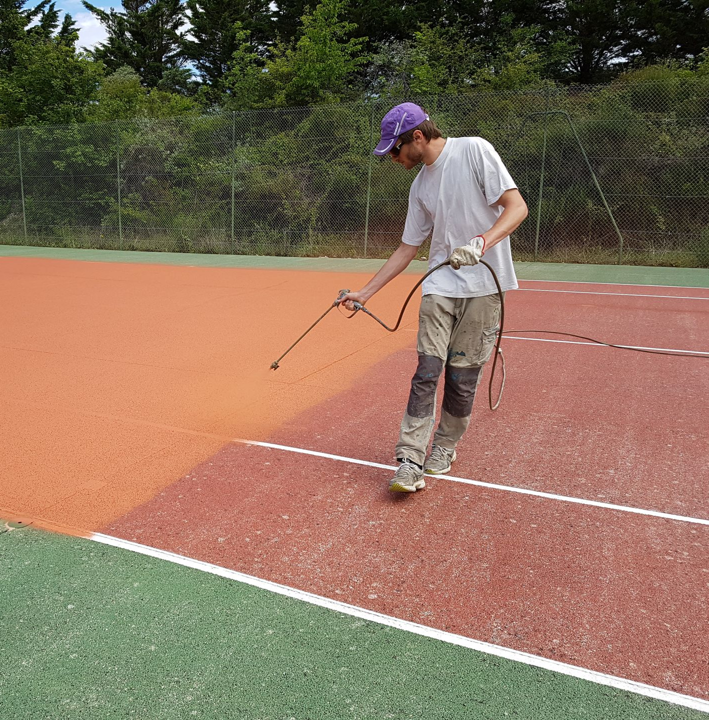 Un homme vaporise de la peinture orange sur un court de tennis, portant un chapeau violet et des genouillères.