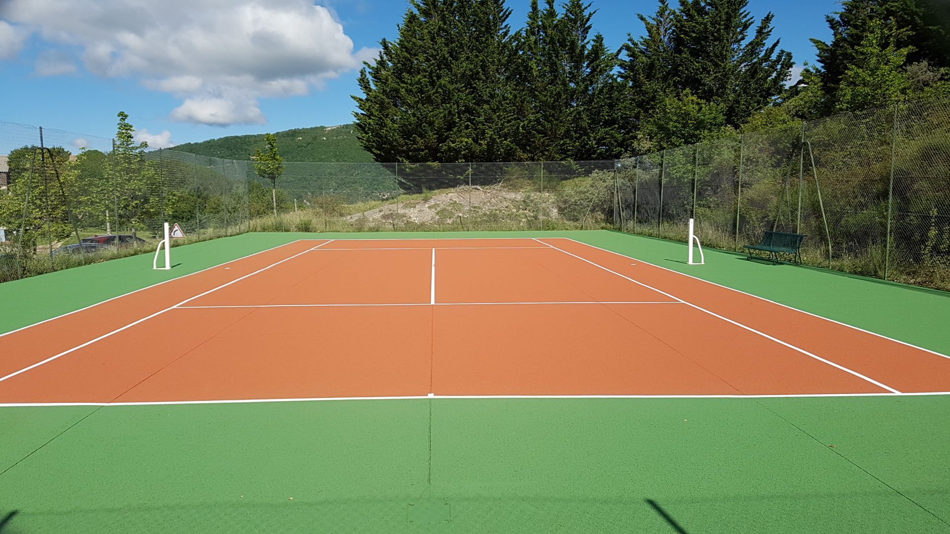 Court de tennis en terre battue rouge entouré de gazon synthétique vert, sous un ciel bleu.