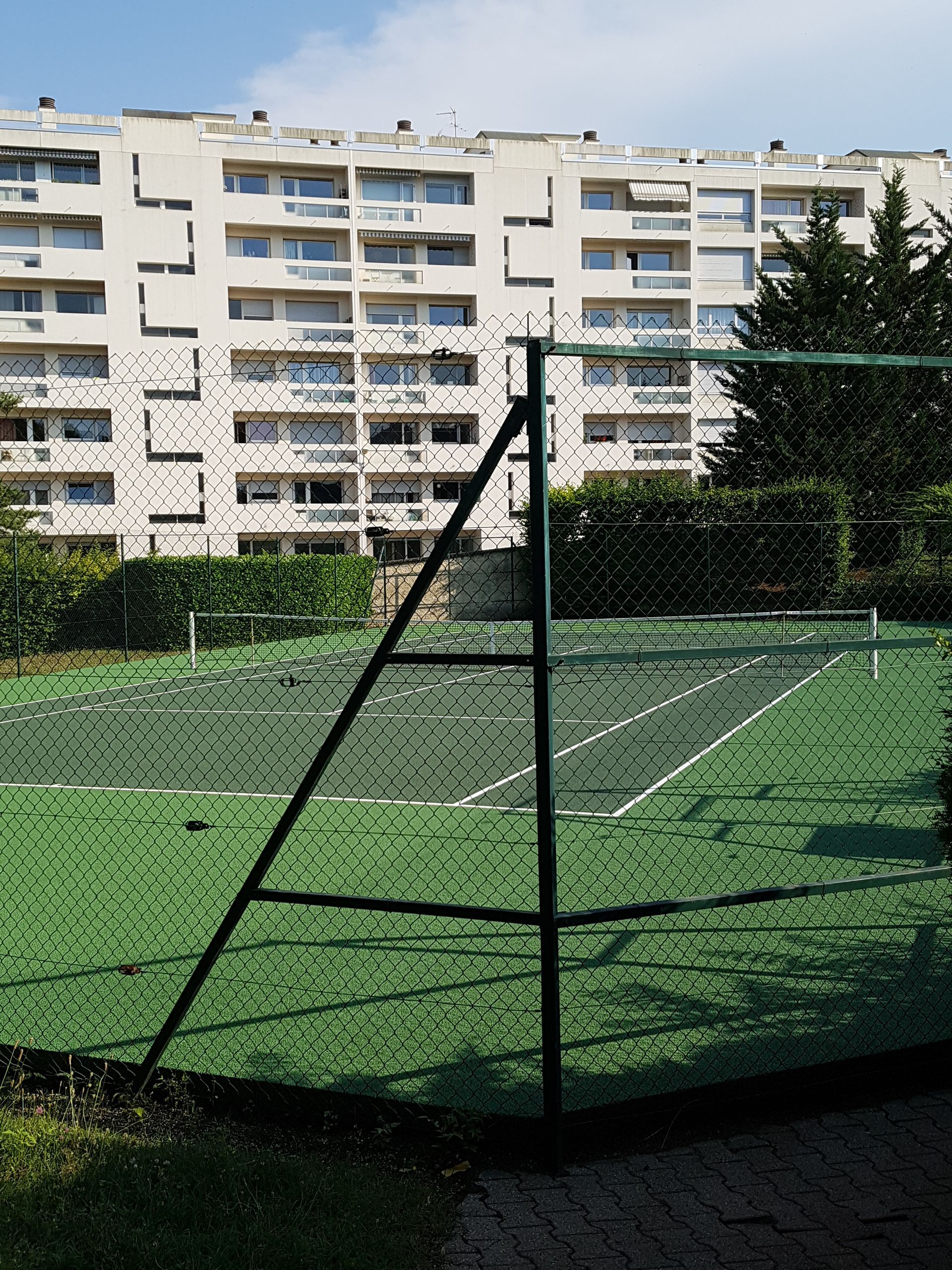Court de tennis à surface verte, clôture noire et immeuble d'appartements en arrière-plan. Journée ensoleillée.