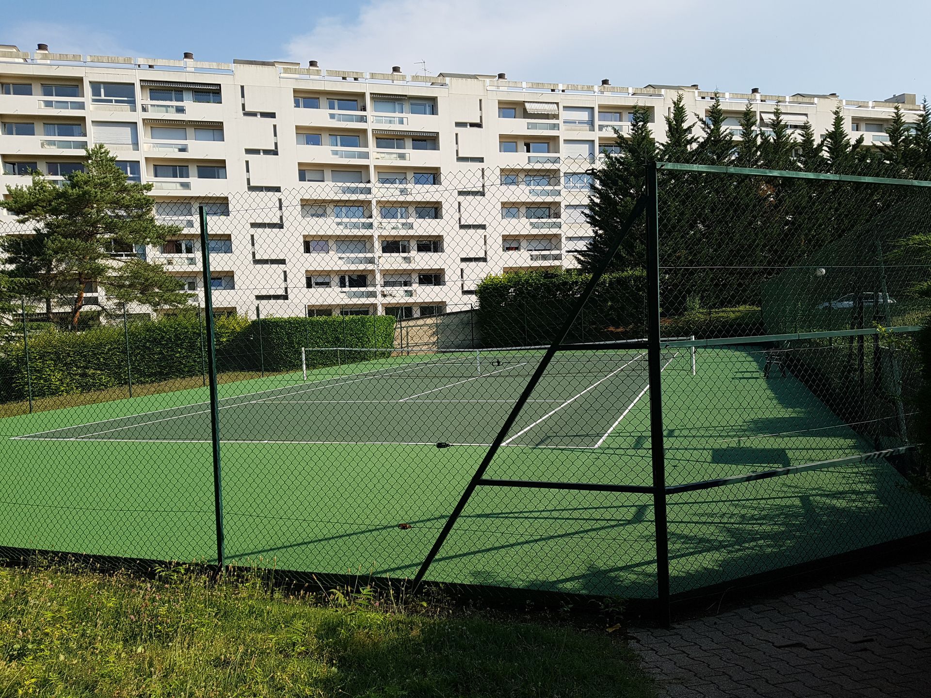 Court de tennis à surface verte, entouré d'une clôture verte et de feuillage, avec un grand immeuble d'appartements en arrière-plan.