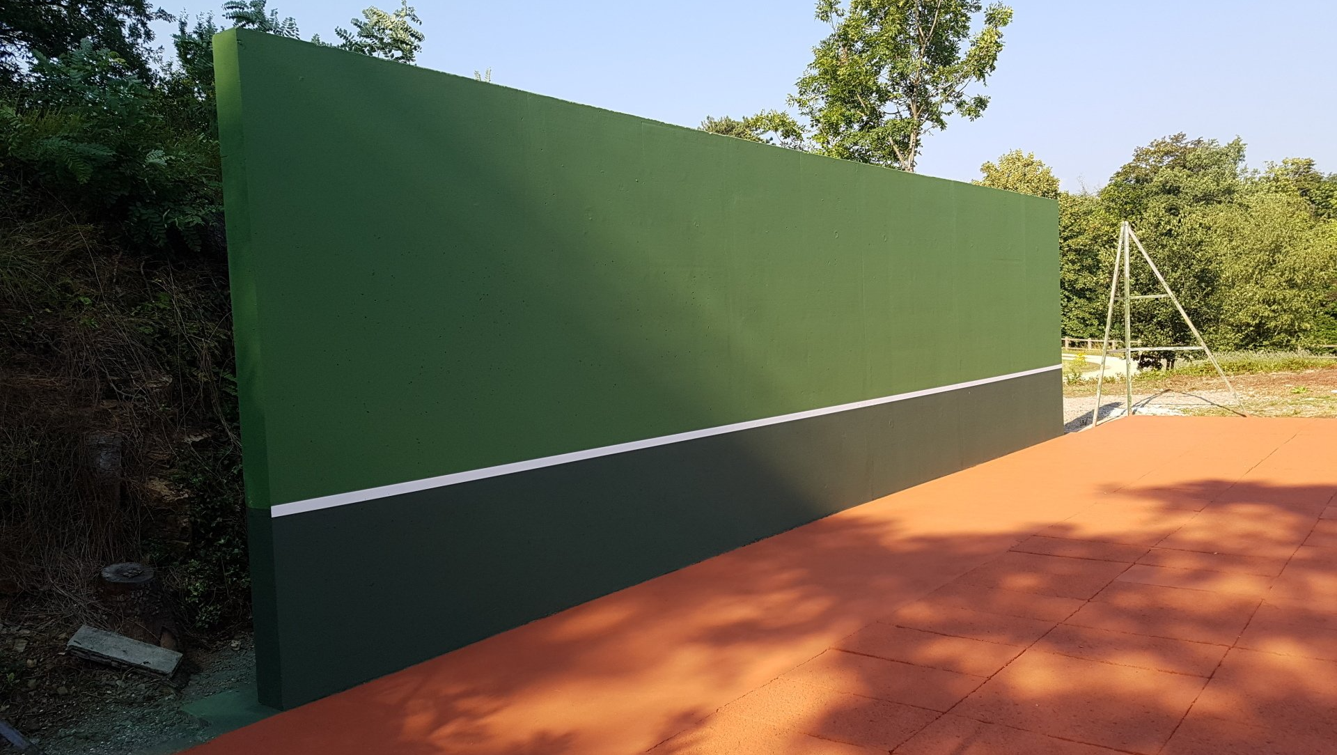 Mur d'entraînement de tennis vert et noir avec une ligne blanche, installé sur un court en terre battue rouge.