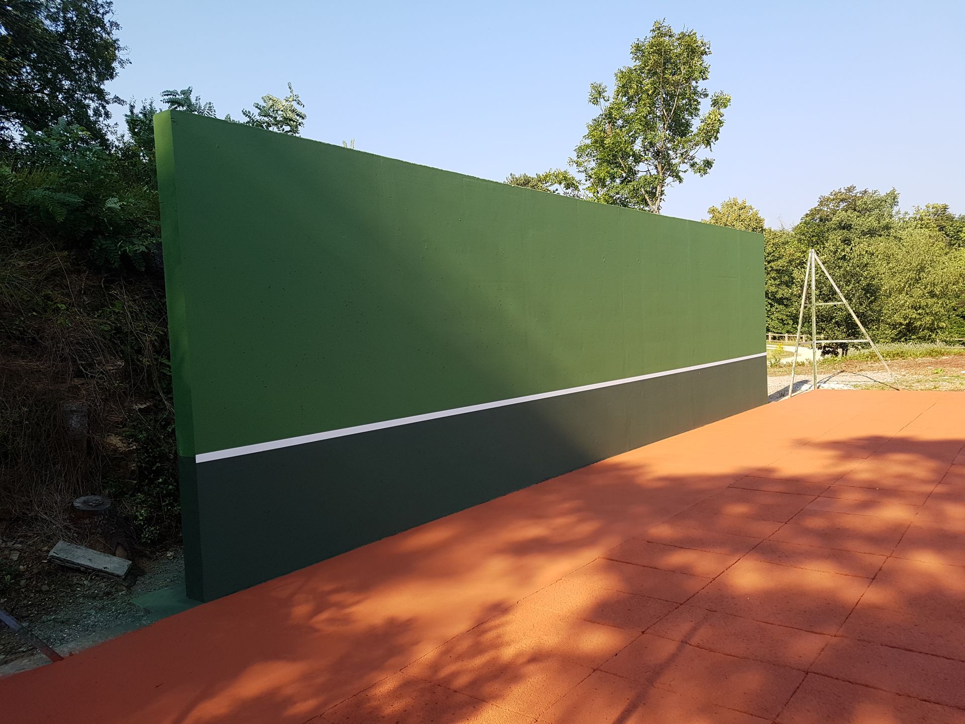 Mur d'entraînement de tennis vert et noir avec une ligne blanche, installé sur un court rouge, avec des arbres en arrière-plan.