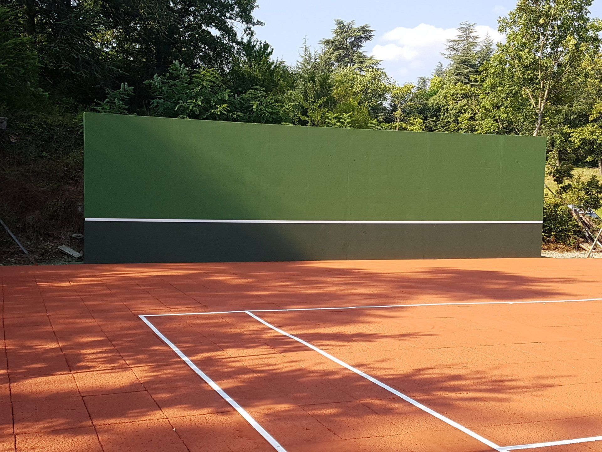 Mur d'entraînement de tennis avec panneaux verts et vert foncé, ligne blanche et court en terre battue. Arbres en arrière-plan.