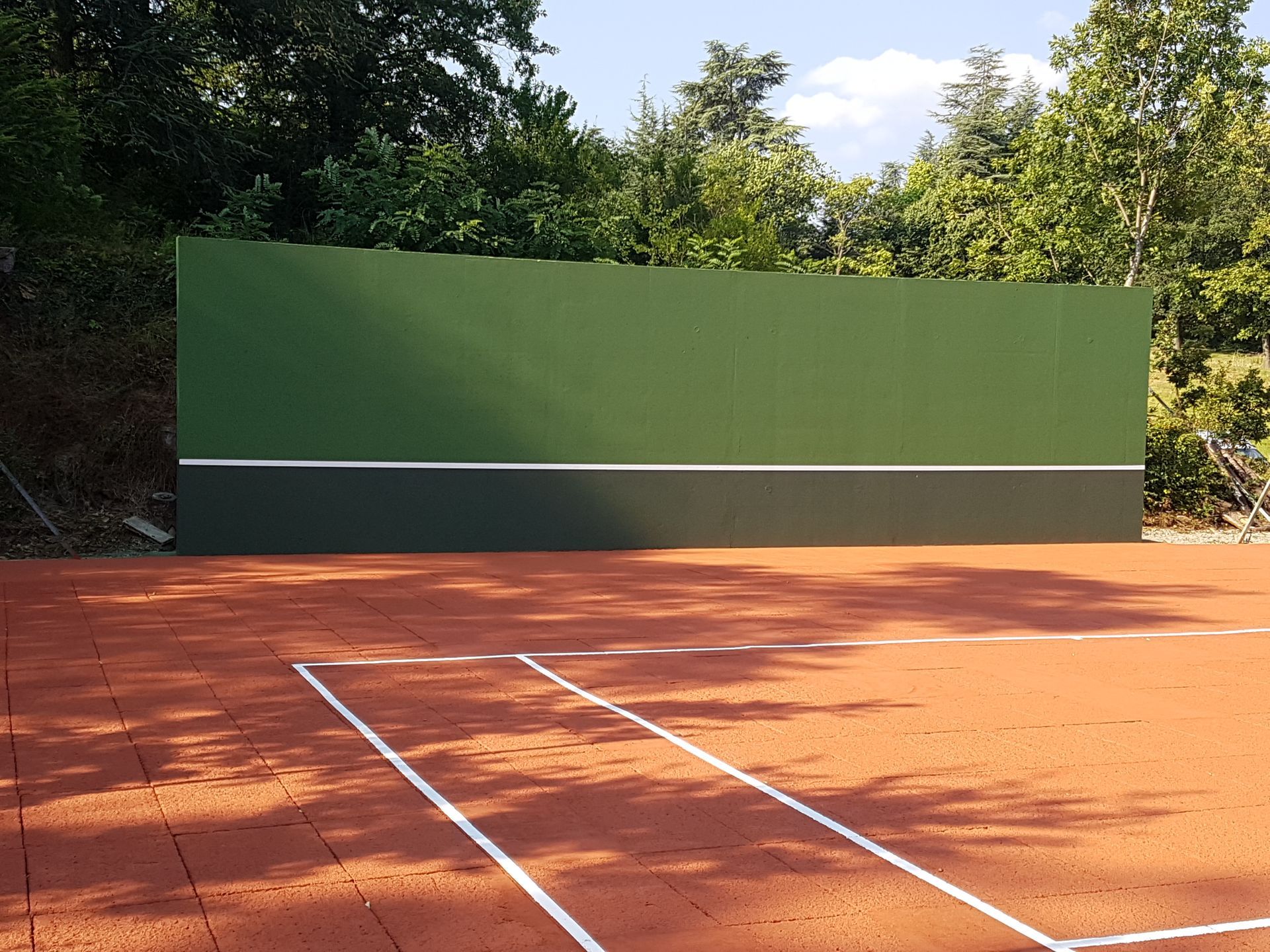 Mur d'entraînement vert et noir sur un court de tennis en terre battue rouge avec des lignes blanches, arbres en arrière-plan.