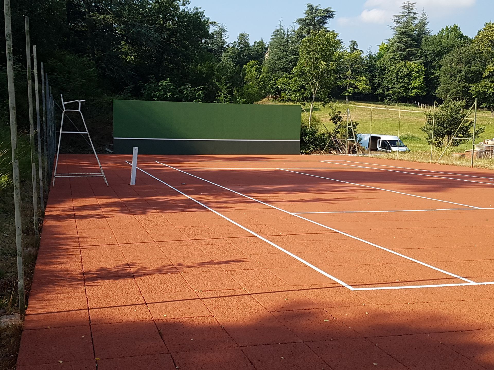 Court de tennis avec surface rouge et mur vert, entouré d'arbres.