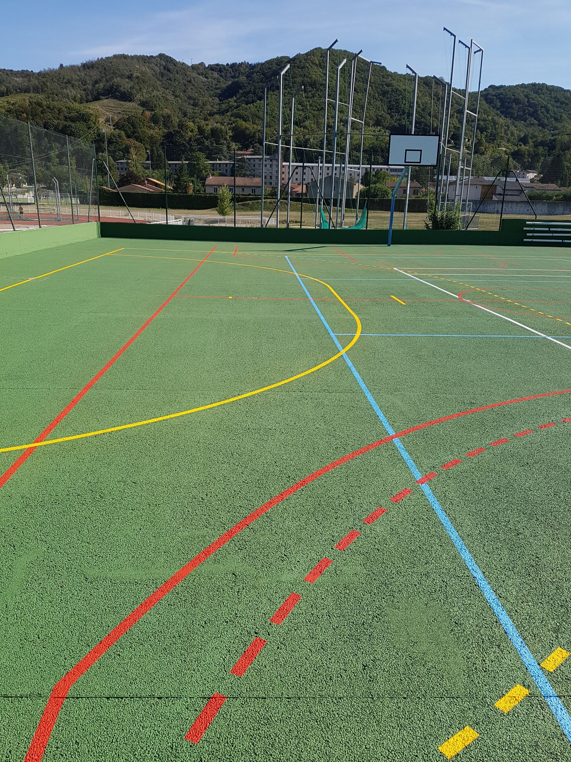 Terrain de sport en gazon synthétique avec panier de basket et lignes colorées, montagnes en arrière-plan.