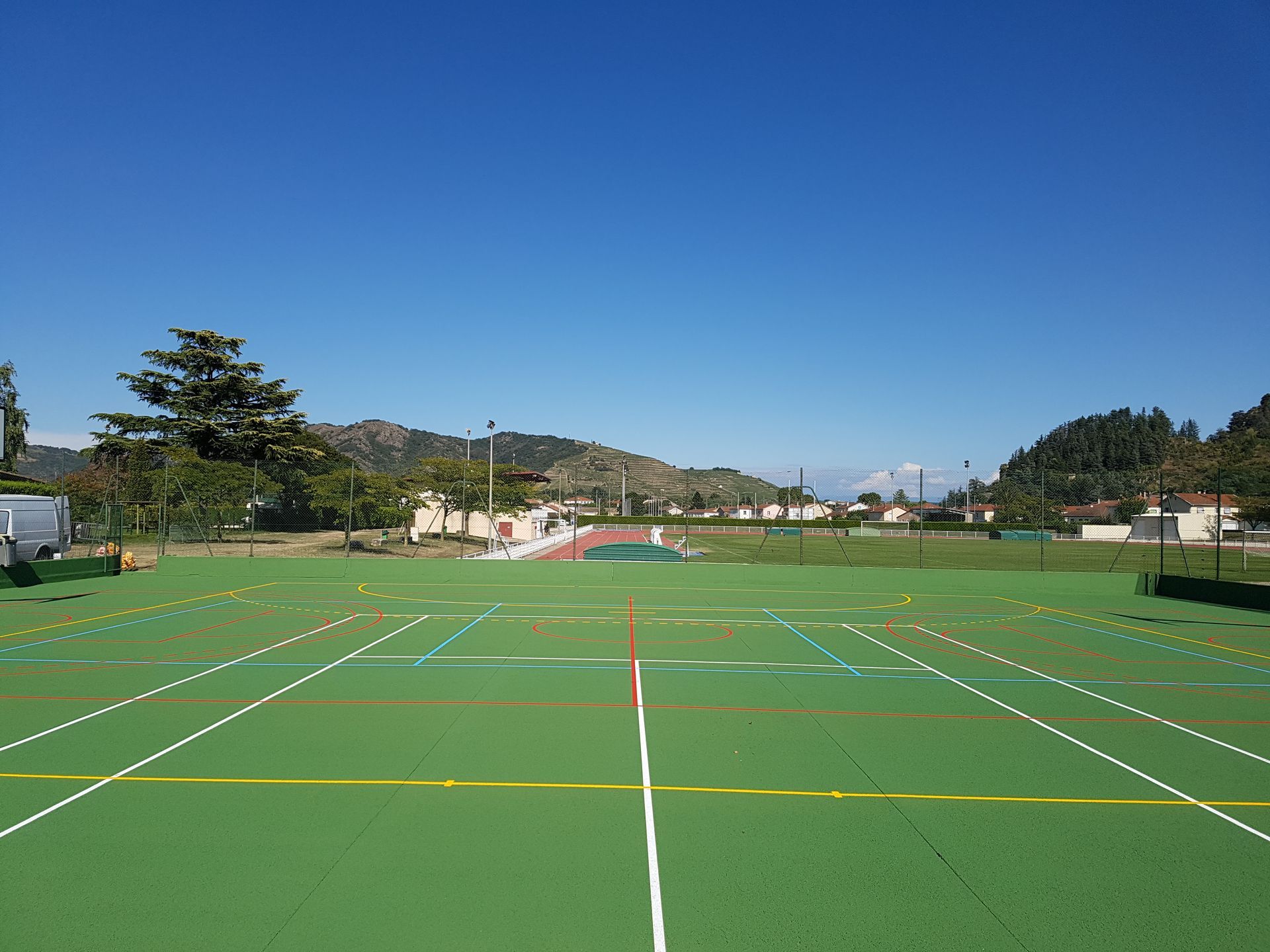 Terrain de sport en gazon synthétique avec lignes peintes, ciel bleu et bâtiments au loin.