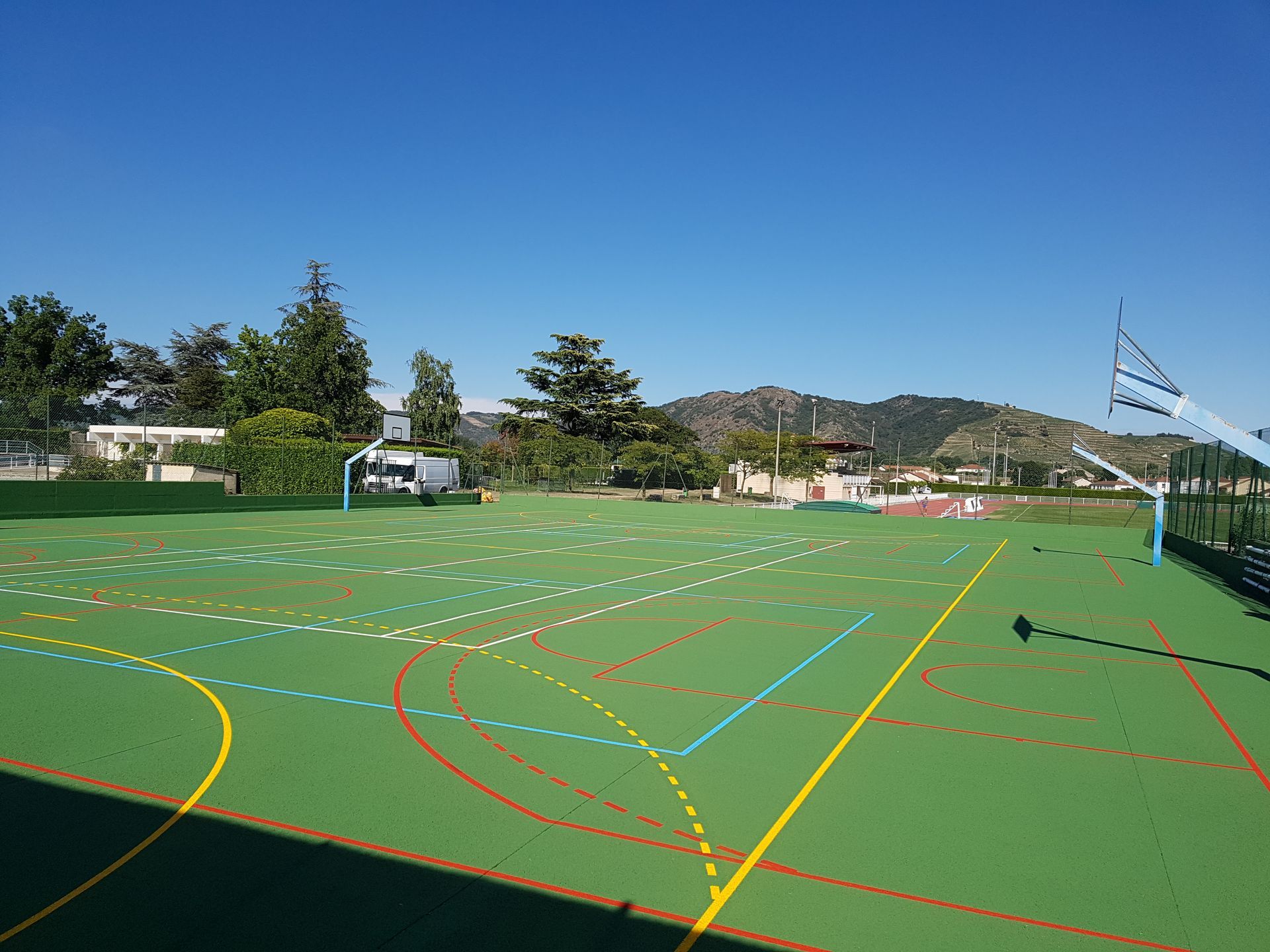 Terrain de sport avec gazon synthétique, lignes peintes et un panier de basket, sous un ciel bleu.