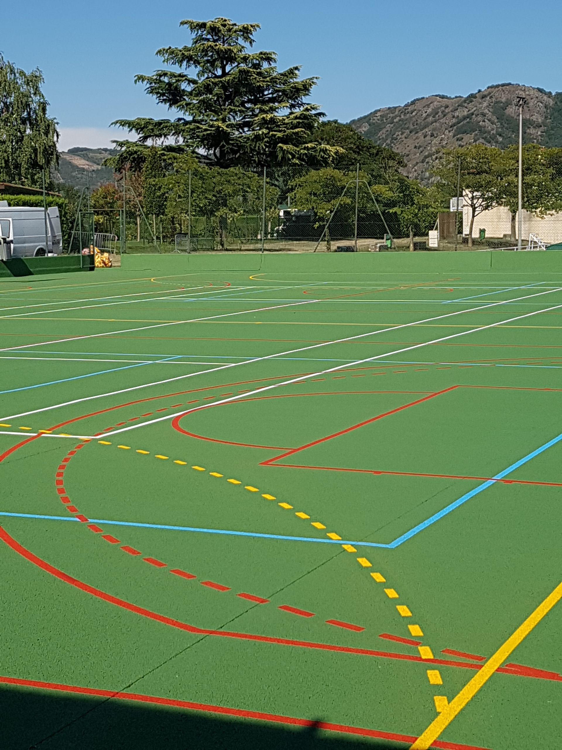 Terrain de sport vert avec des lignes colorées, des arbres et des montagnes en arrière-plan, sous un ciel bleu.