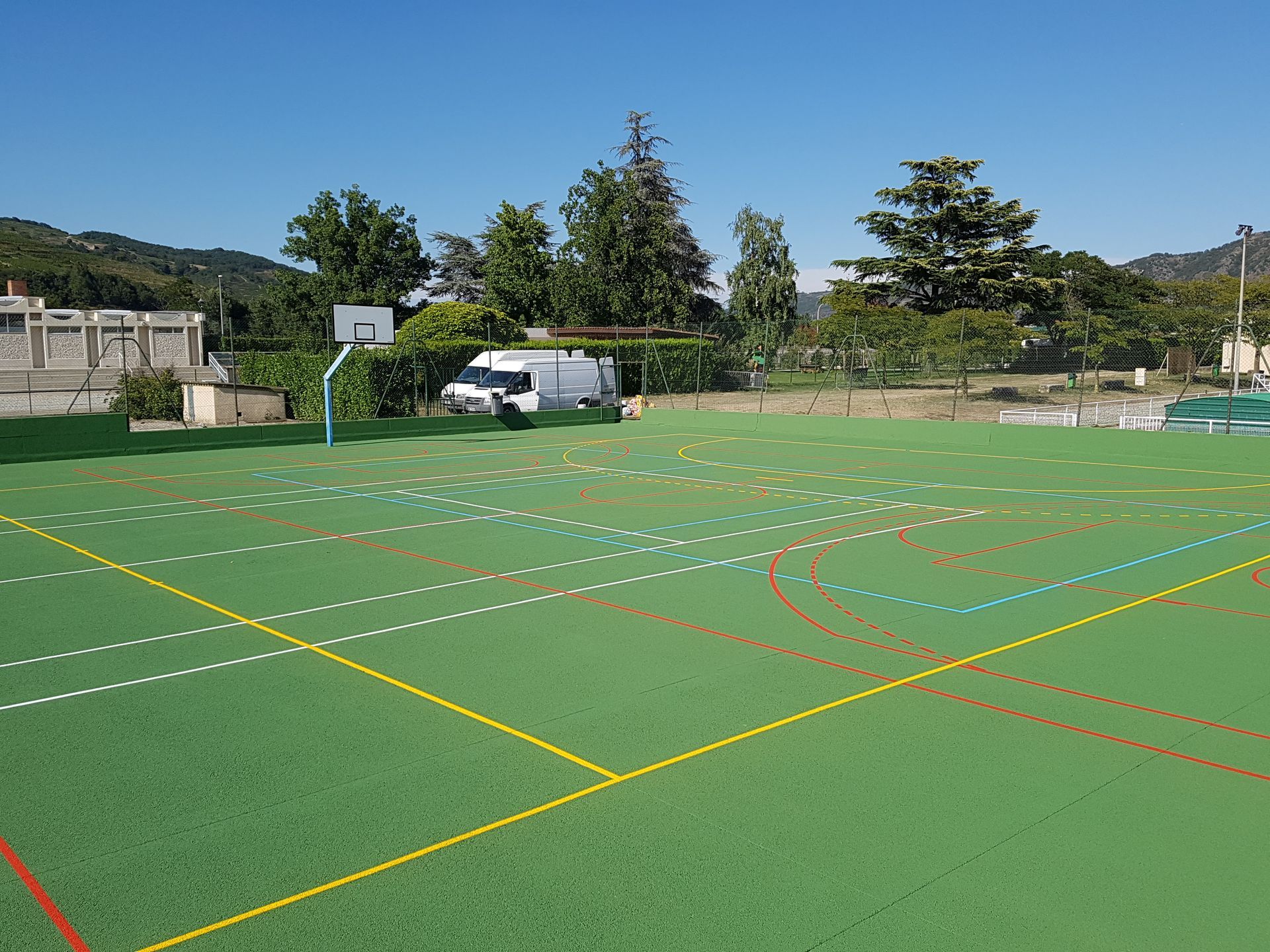Terrain de sport vert avec des lignes blanches, jaunes et rouges, un panier de basket et des arbres sous un ciel bleu.