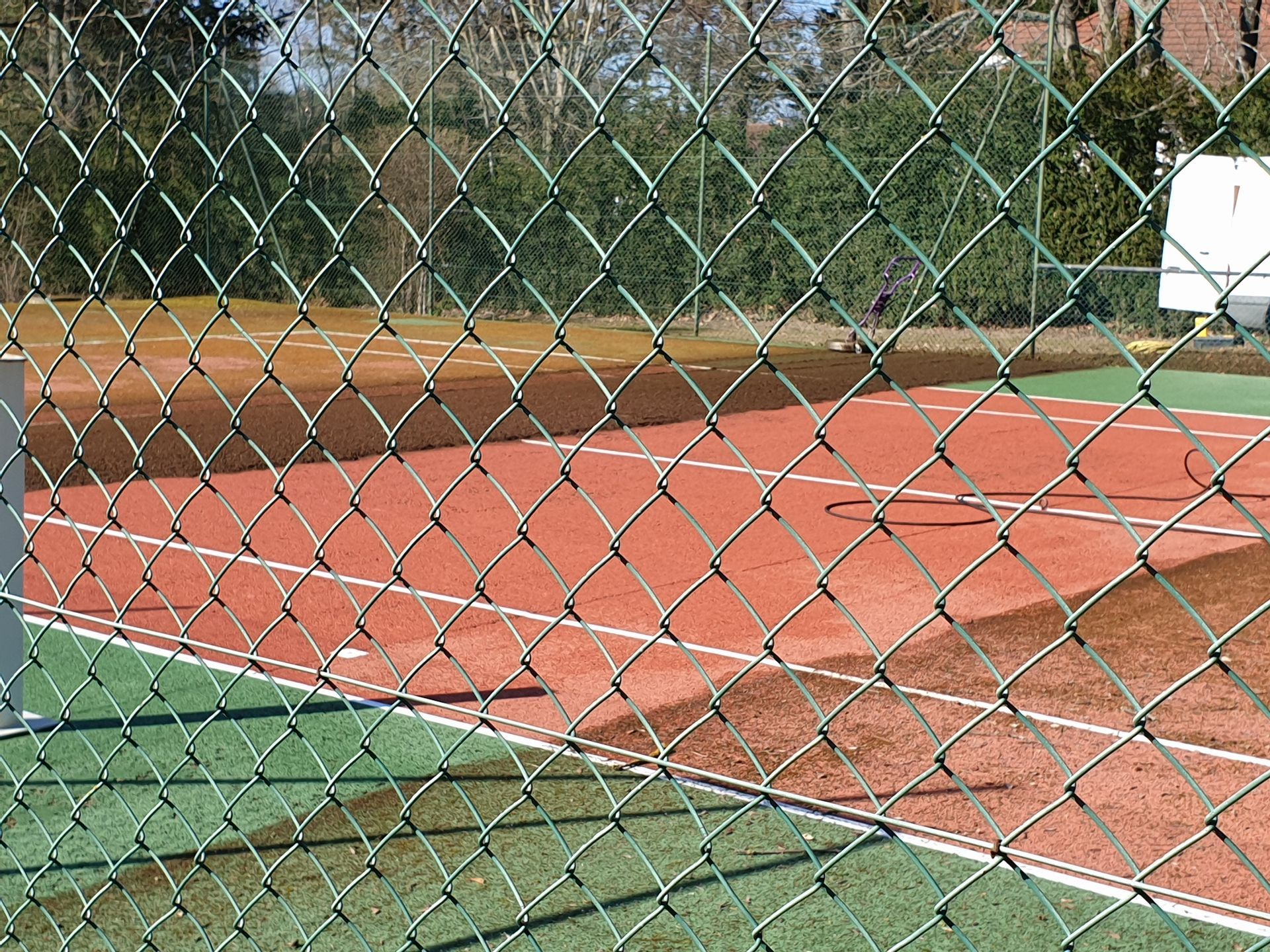 Court de tennis derrière une clôture en grillage ; surface de jeu rouge avec des lignes blanches, zone environnante verte.