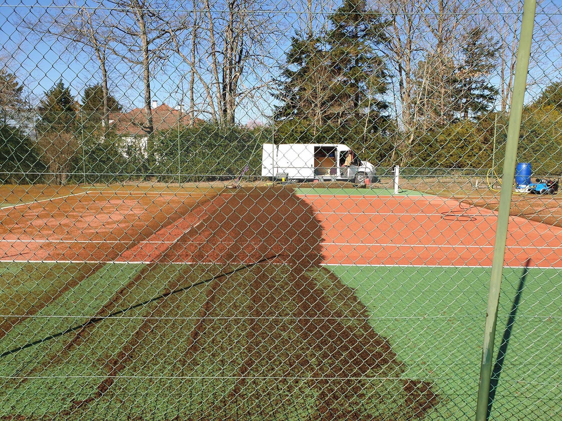 Court de tennis en cours d'entretien ; certaines parties du court rouge et vert sont recouvertes de terre, clôture au premier plan, camion de service à l'arrière-plan.