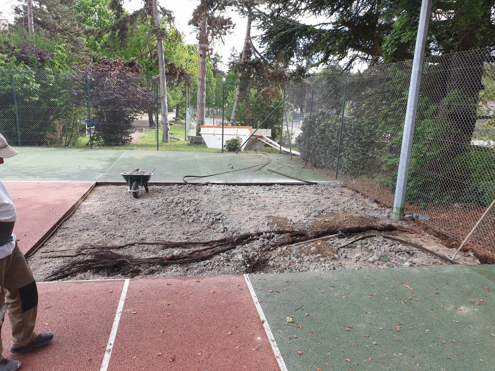 Rénovation d'un court de tennis : zone de gravier, personne avec des outils, filets, arbres en arrière-plan.