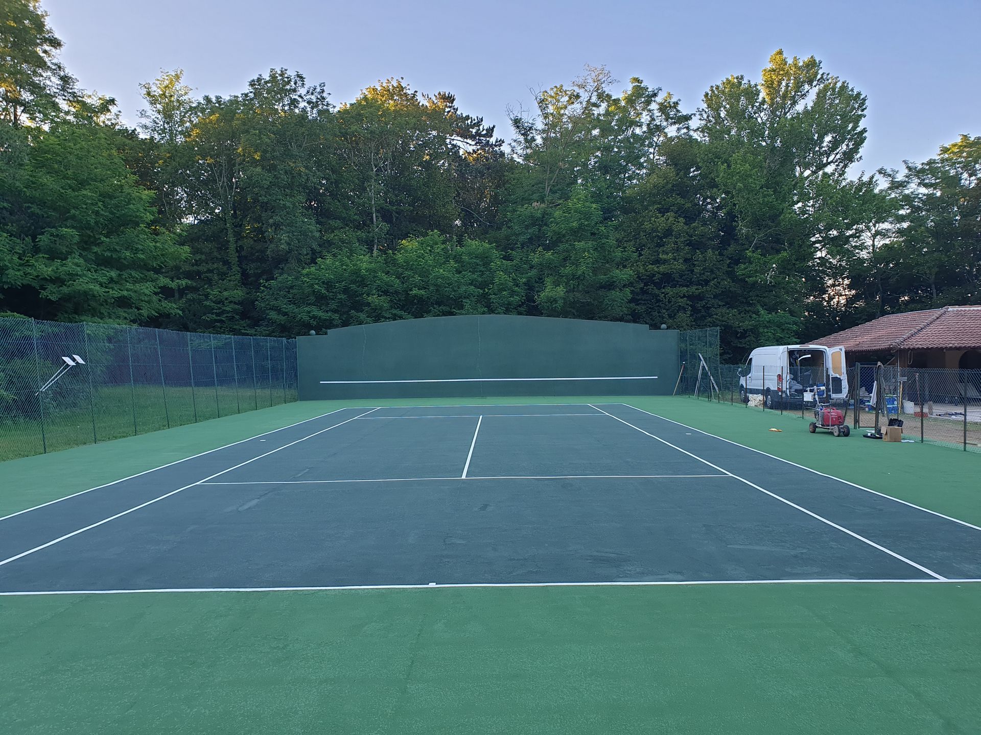 Court de tennis avec surface verte, lignes blanches, fond vert et arbres.