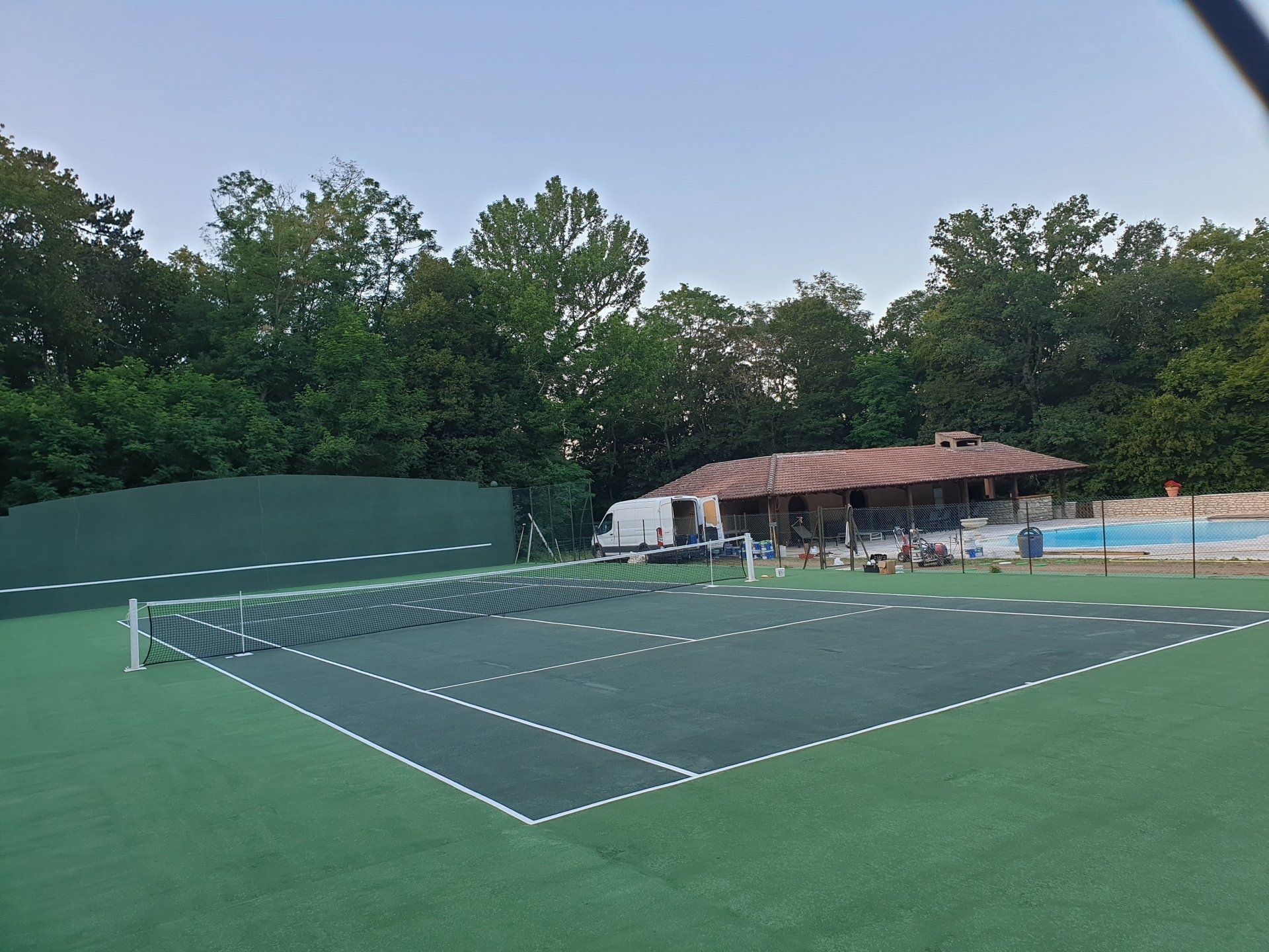 Court de tennis avec surface verte, filet et bâtiment en arrière-plan. Des arbres et une piscine sont visibles.