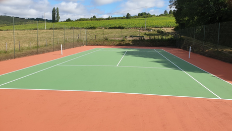Court de tennis, surface de jeu verte, entourée d'orange, avec un champ et des arbres en arrière-plan.