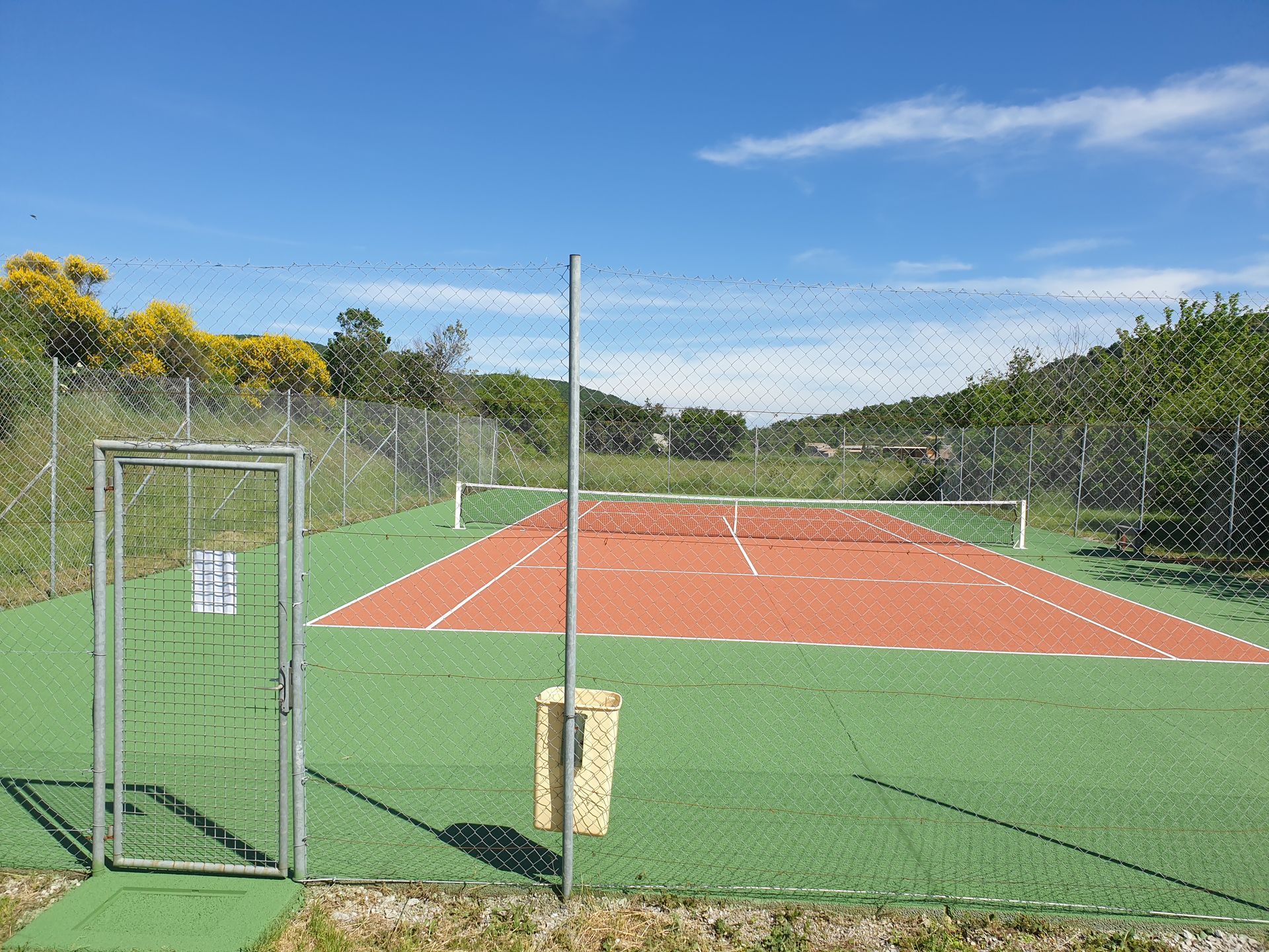 Terrain de sport extérieur clôturé avec surface rouge et verte, filet, portail et en arrière-plan des arbres et un ciel bleu.